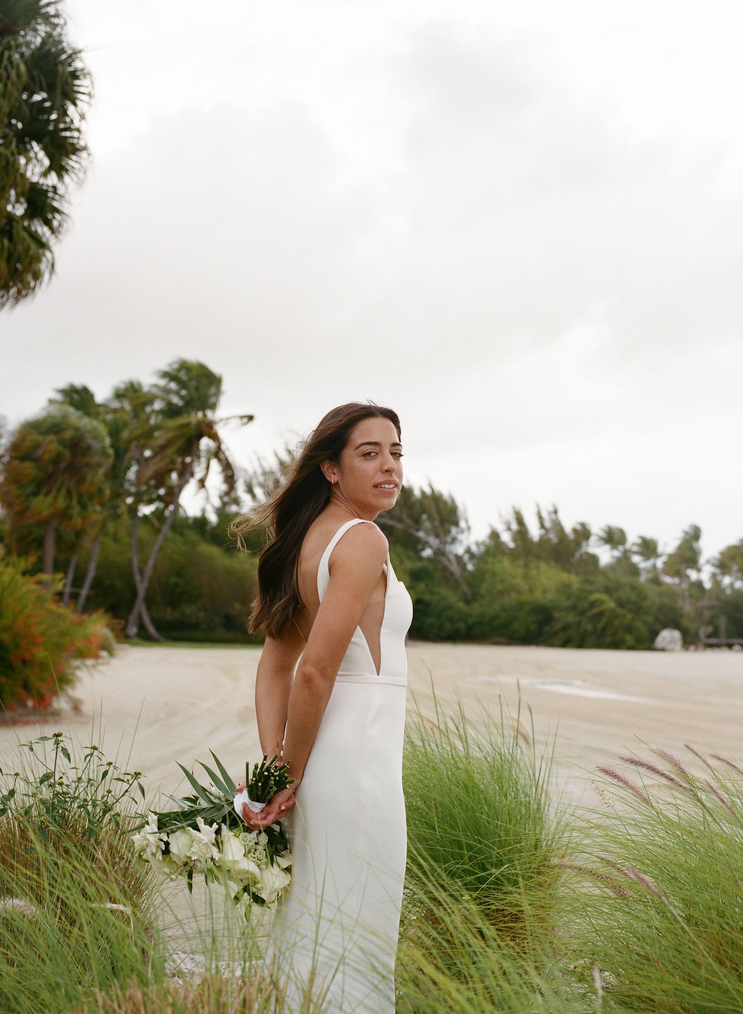 Bride in white gown holding bouquet on tropical beach, palm trees swaying in breeze, overcast sky.