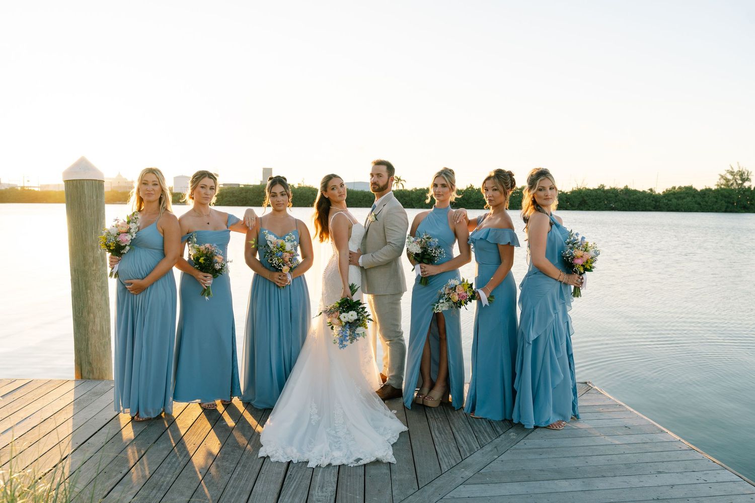 Wedding party in light blue bridesmaid dresses poses together on wooden dock by serene waterfront at sunset.