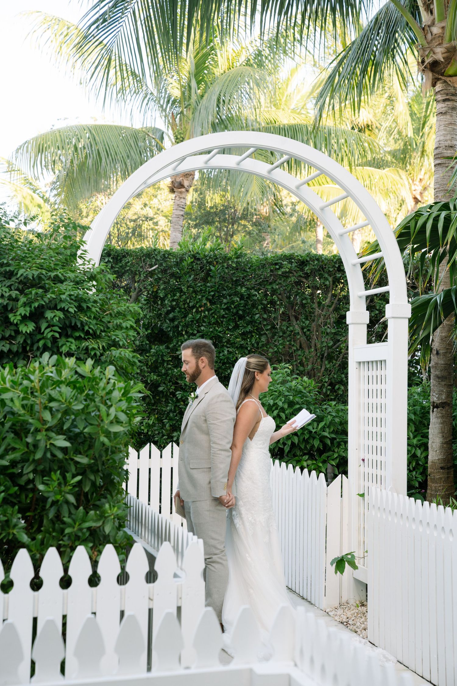 A couple shares an intimate moment under a white garden arbor surrounded by lush palm trees and tropical foliage.