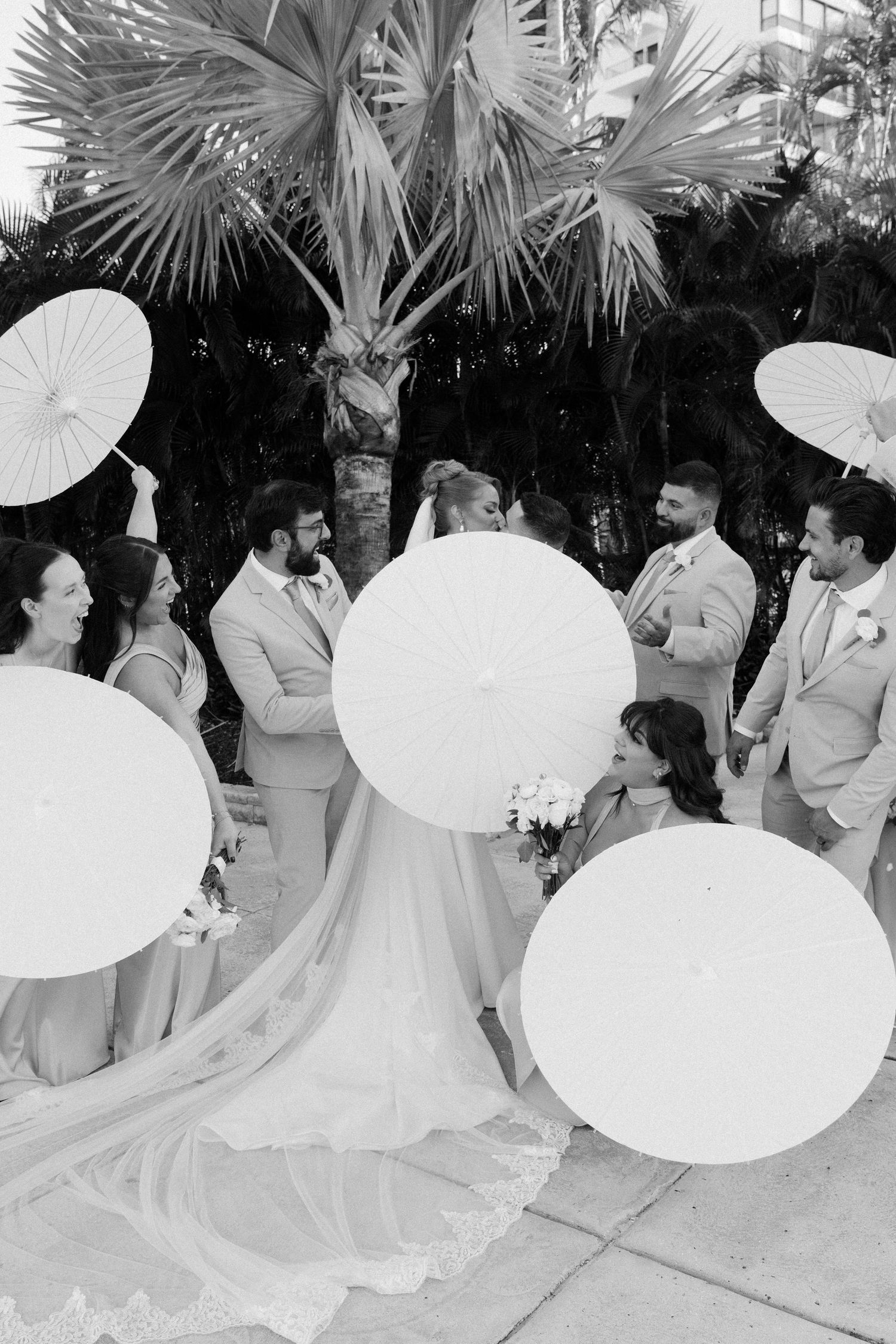 Wedding party poses with decorative white parasols among tropical palm trees.
