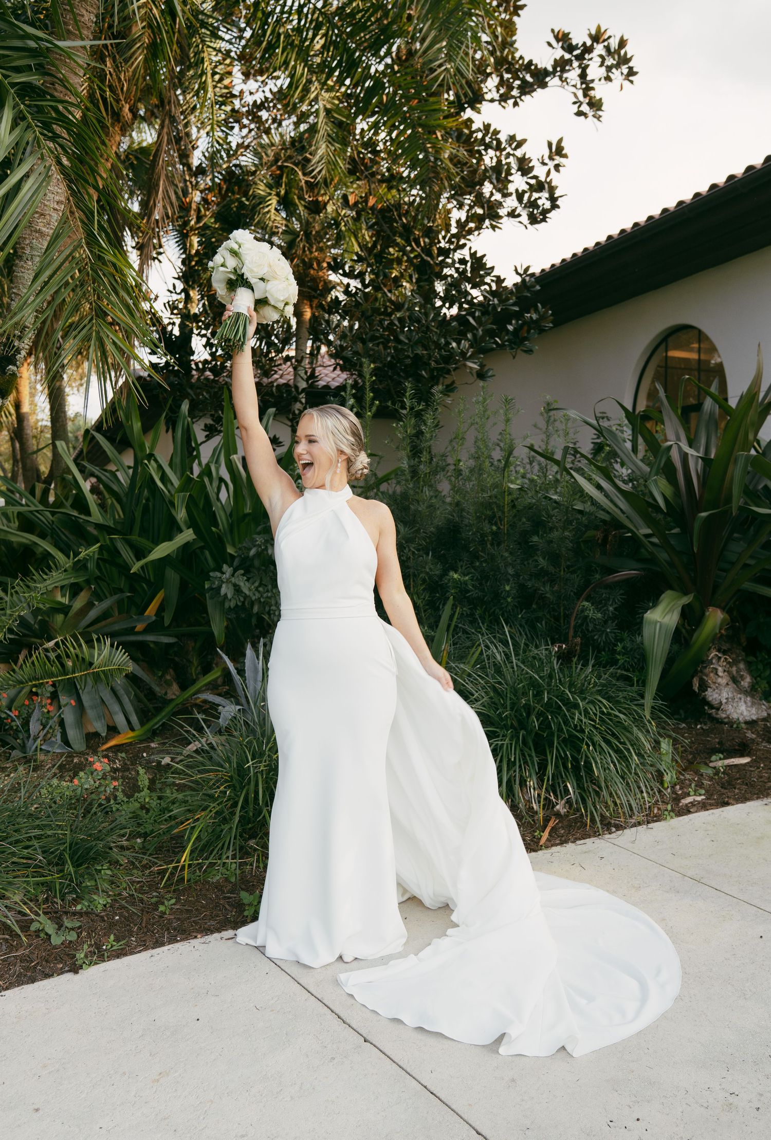 A person in an elegant white wedding gown with a flowing train poses outdoors against a tropical garden backdrop.