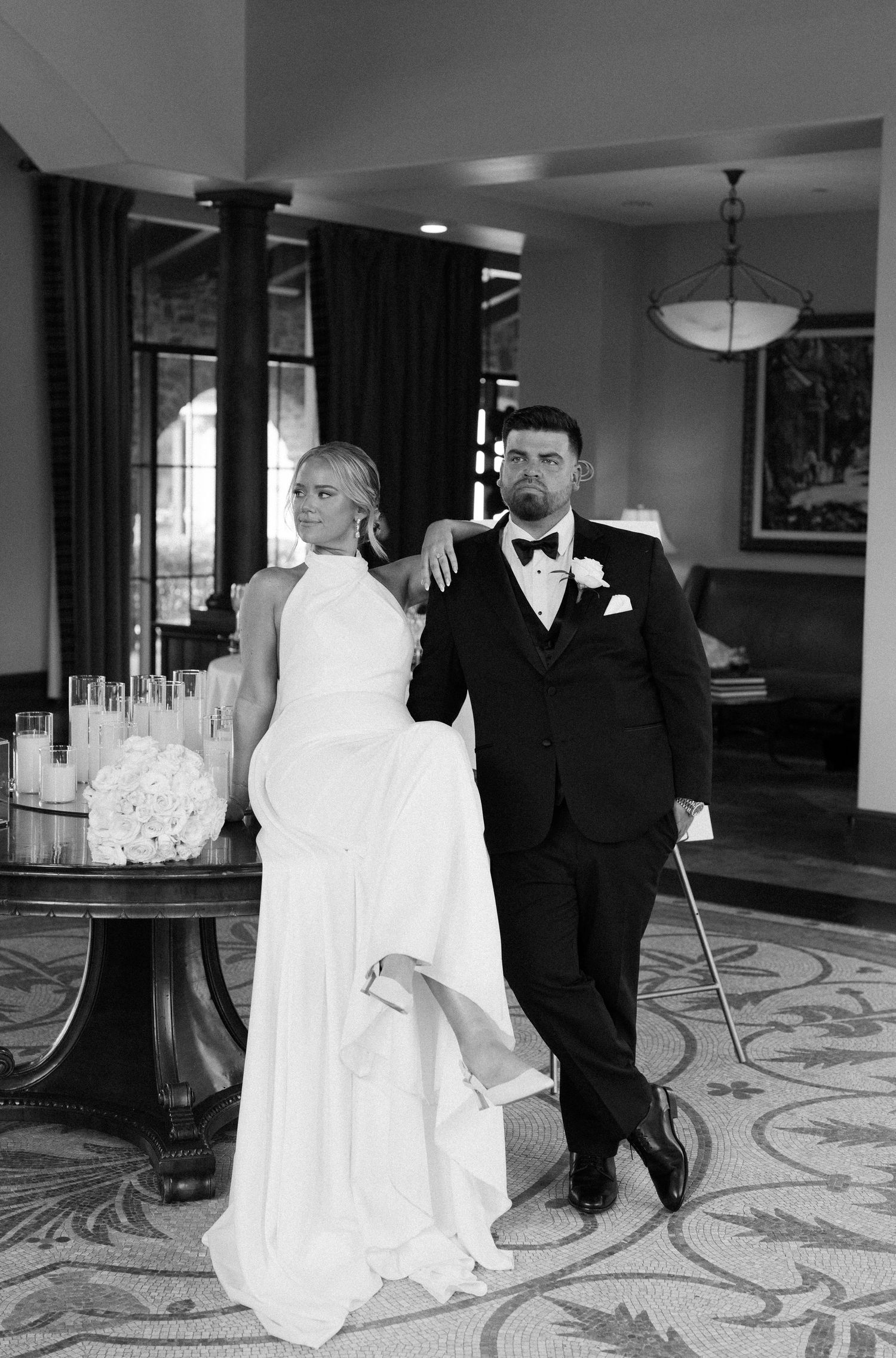 A black and white wedding photo showing a couple in formal attire posing elegantly in a luxurious hotel setting.
