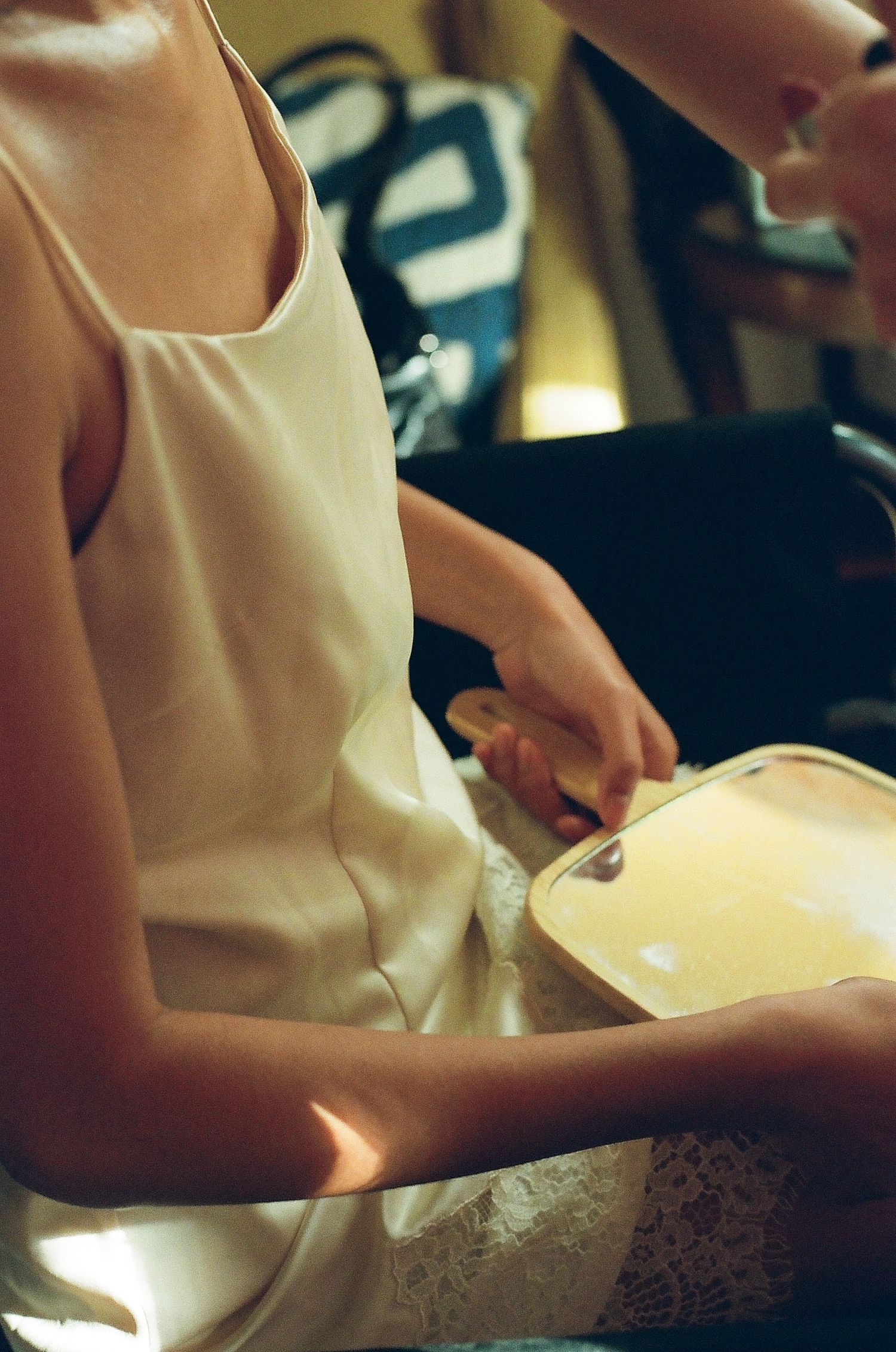 Woman in white camisole holding an open cosmetic case, seated indoors with warm lighting.