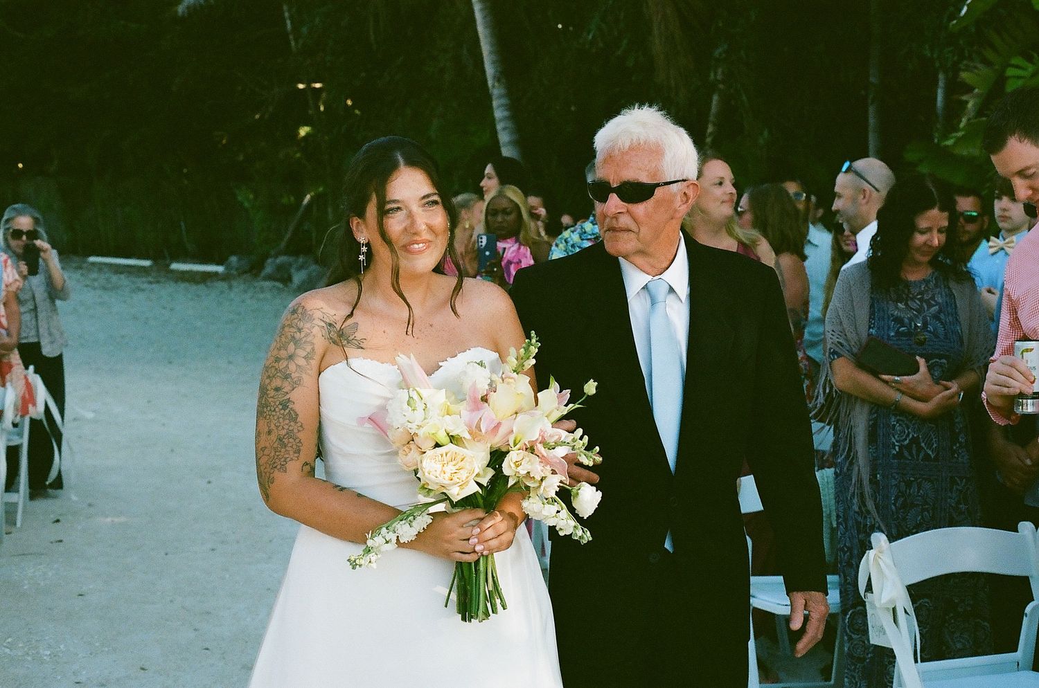 Bride with tattoos walks down outdoor aisle holding bouquet with older man in suit and sunglasses.