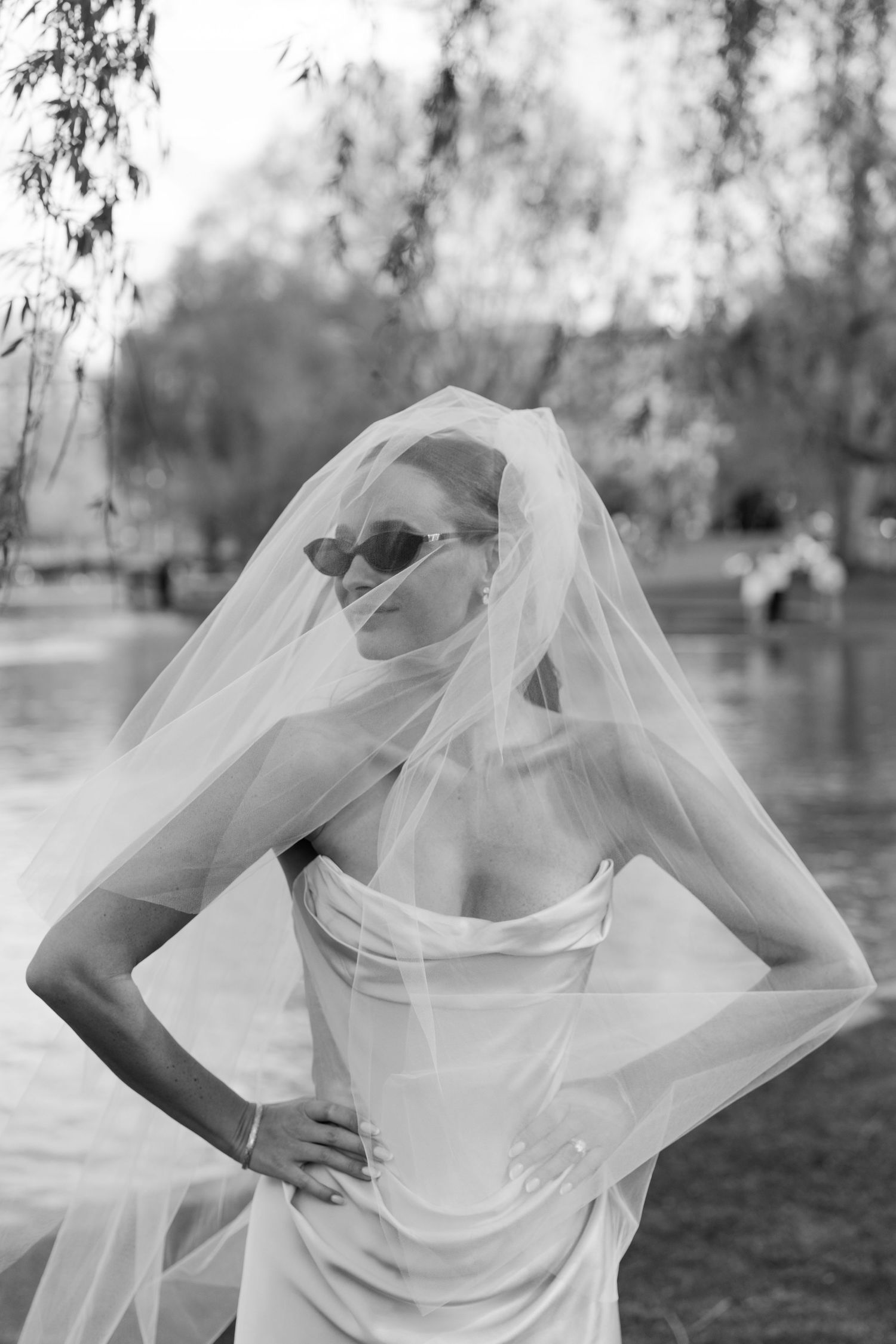 Stylish bride in strapless gown and sunglasses with veil outdoors near a lake in black and white photo.