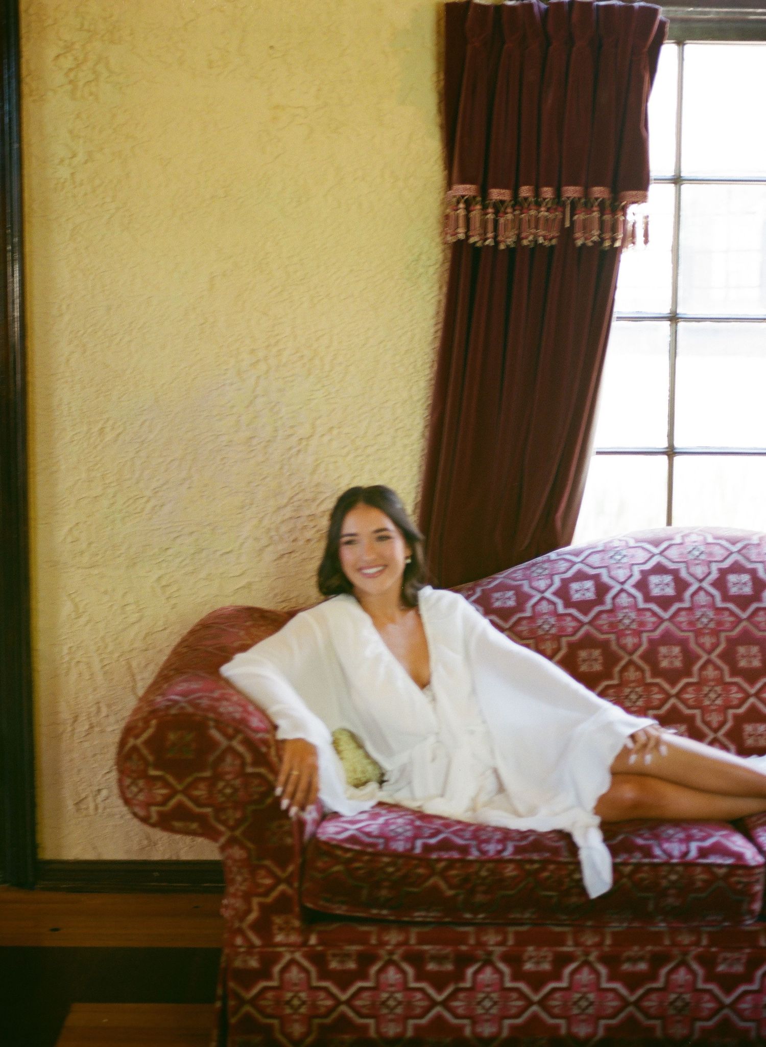 Bride in a white robe relaxes on a vintage red floral sofa near a draped window before her wedding.