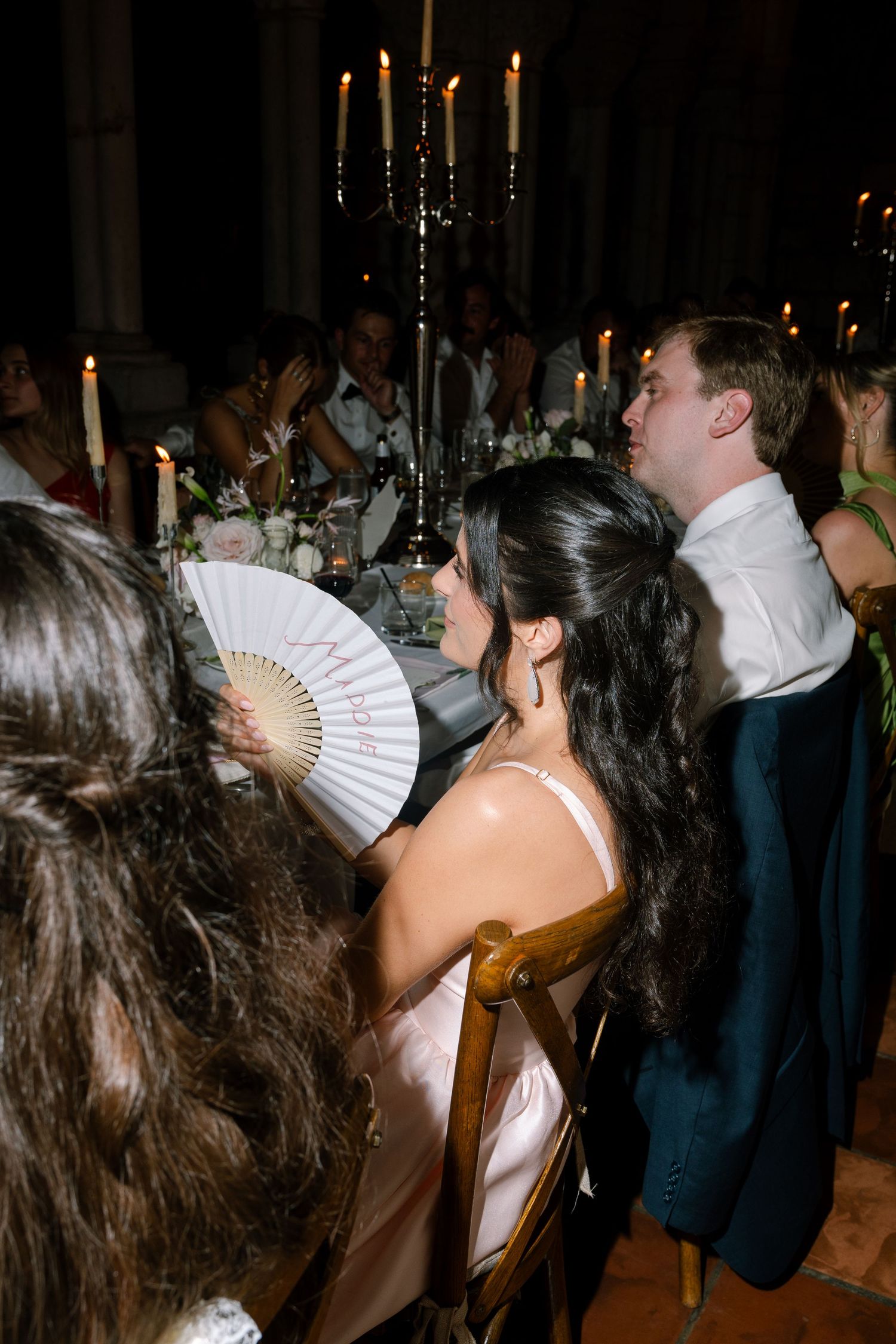Wedding guests seated at candlelit reception table, woman in pink dress holding a decorative white fan.