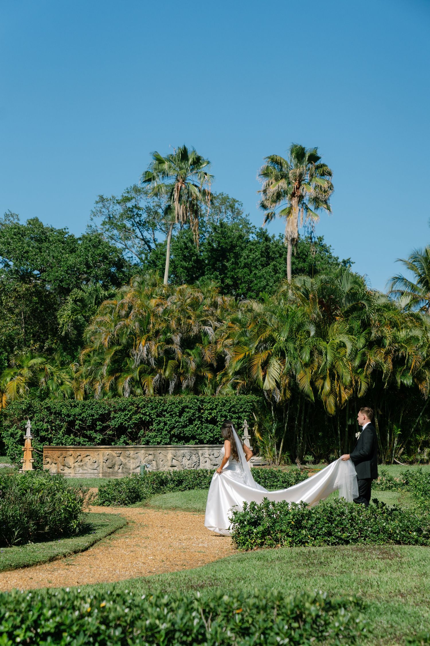 Bride and groom standing in formal garden with palm trees and blue sky on wedding day.