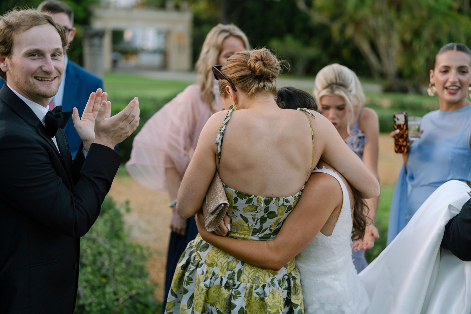 Bride embraces guest in floral dress while man in tuxedo applauds during outdoor wedding reception.
