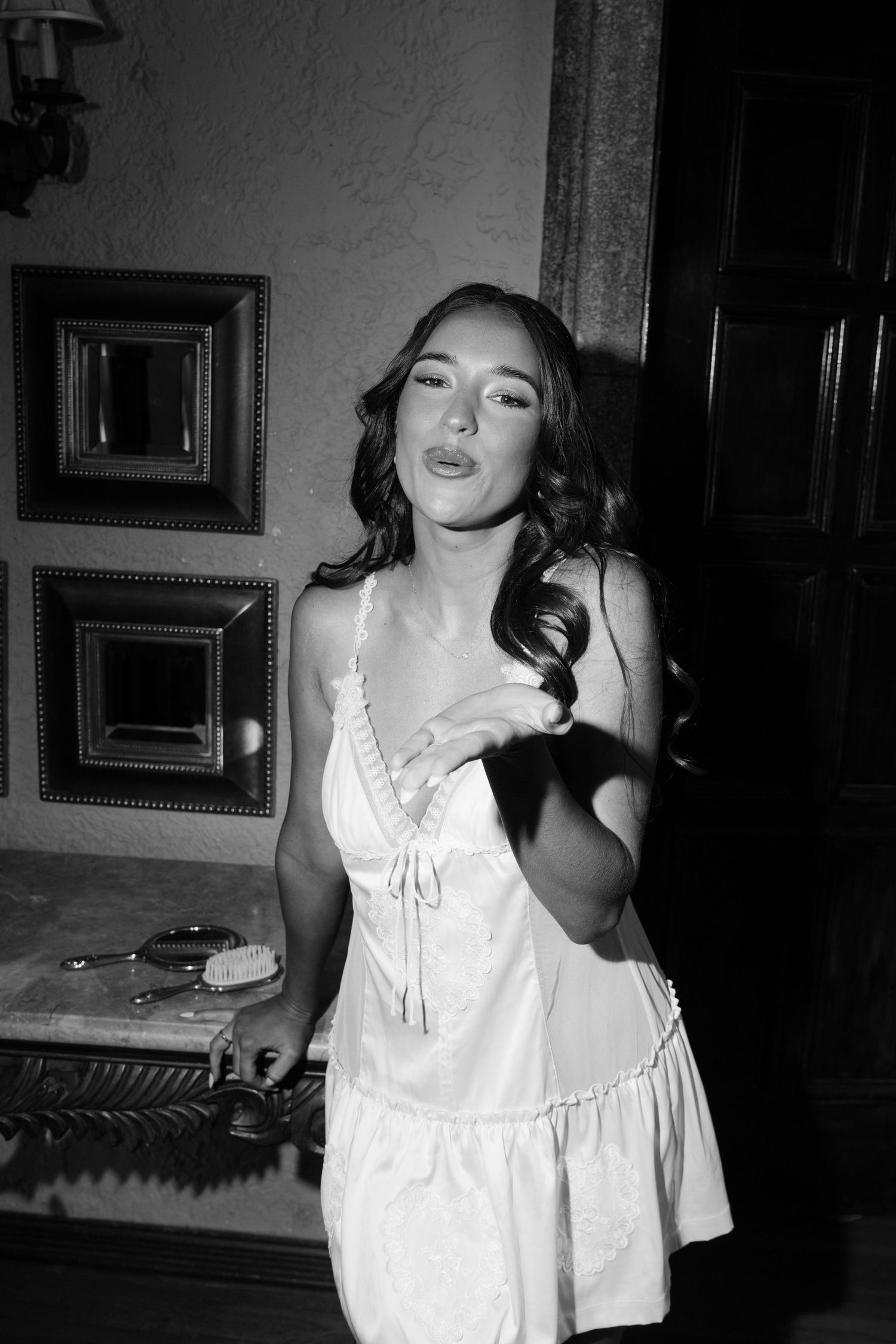 Black and white photo of a young woman in a white slip dress posing indoors near ornate framed mirrors.