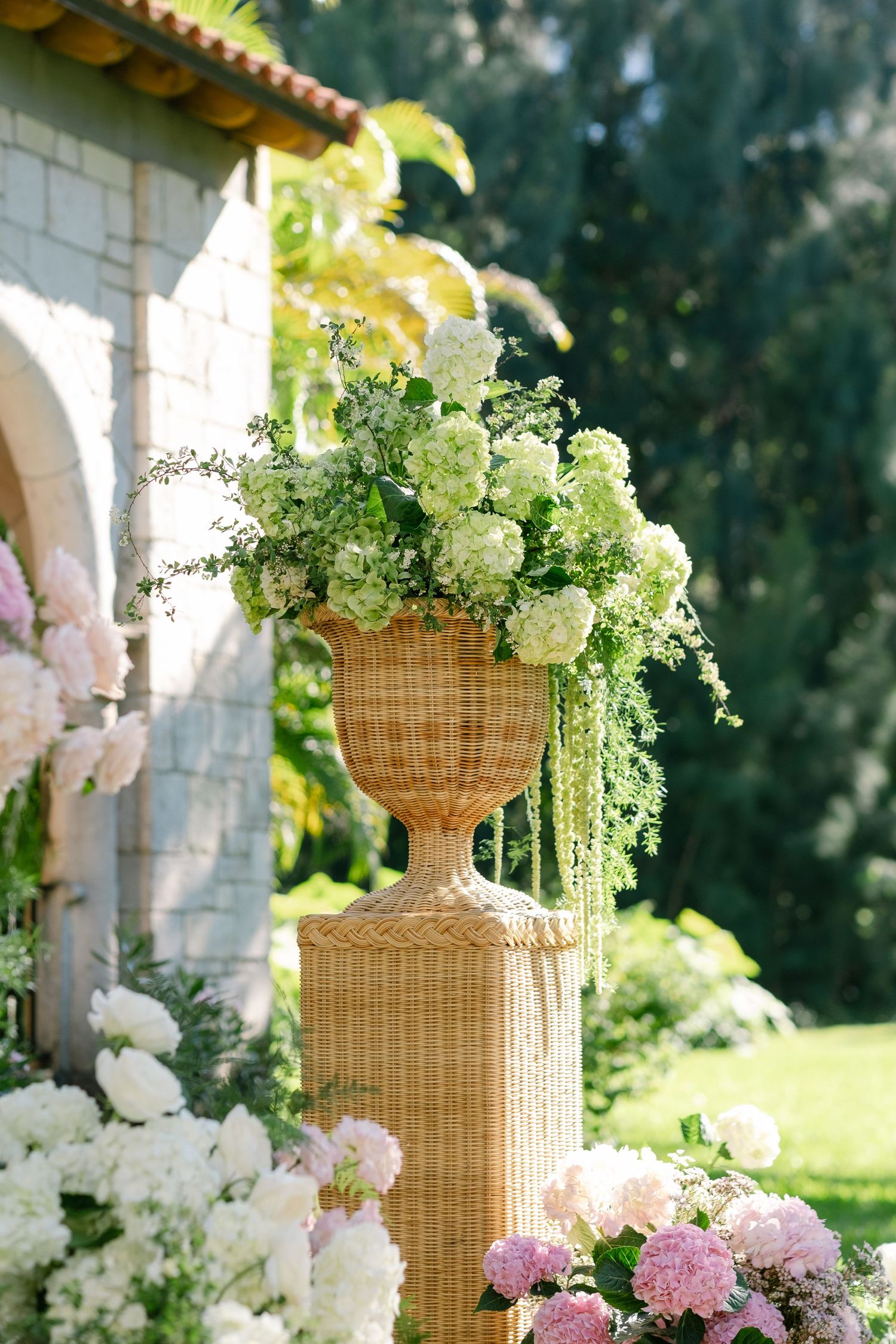 Elegant wicker urn filled with lush white hydrangeas and greenery on a pedestal at an outdoor wedding ceremony.