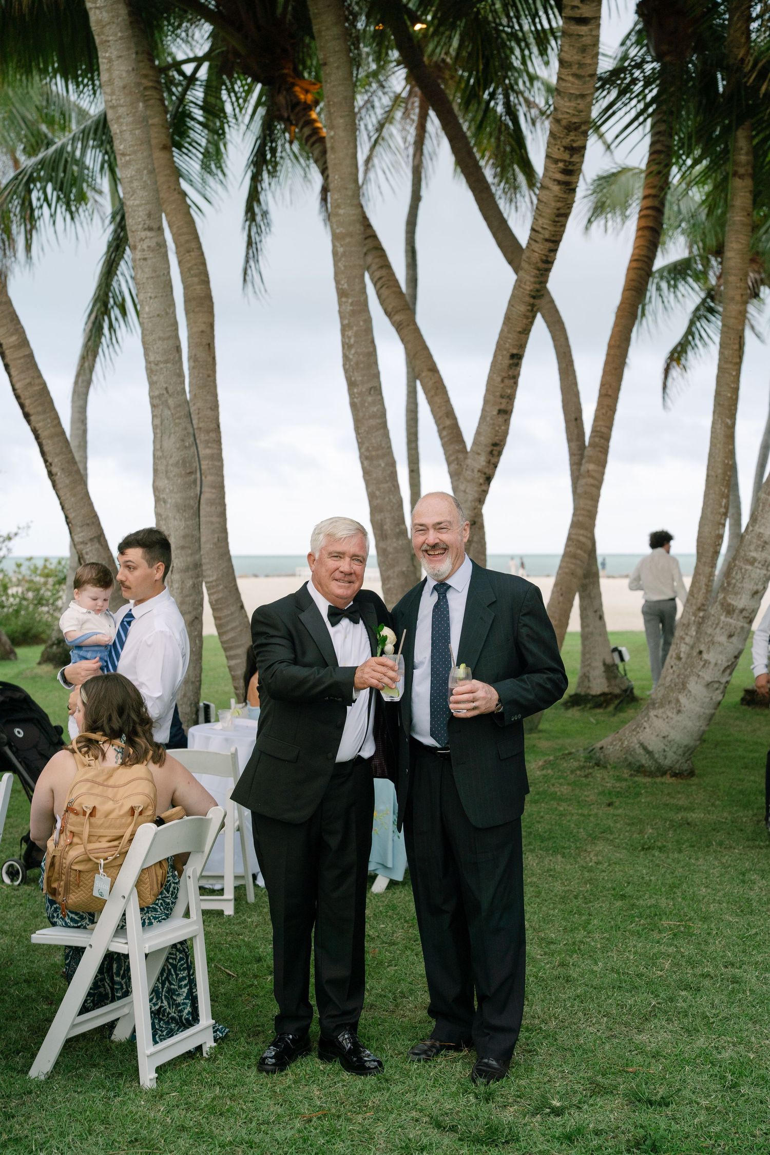Two men in formal tuxedos smile and hold drinks at an outdoor beachside wedding reception with palm trees.