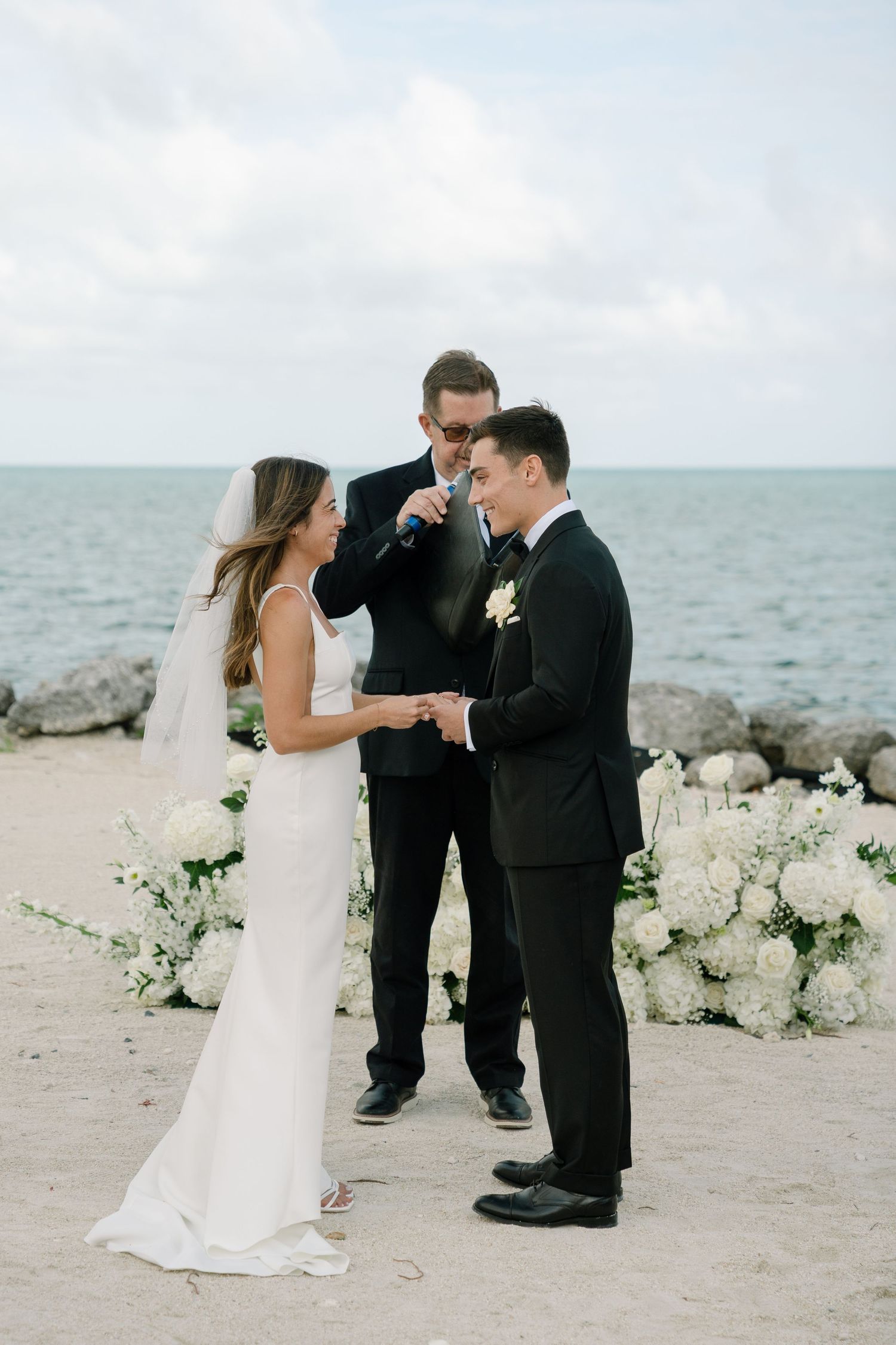 Bride places ring on groom's finger during beachside wedding ceremony with officiant and white floral arrangements.