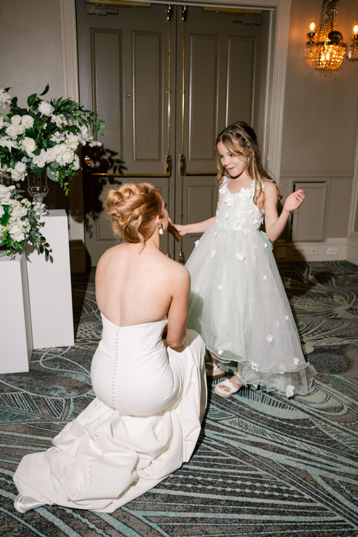 Wedding party members sit on patterned carpet in elegant white dresses.