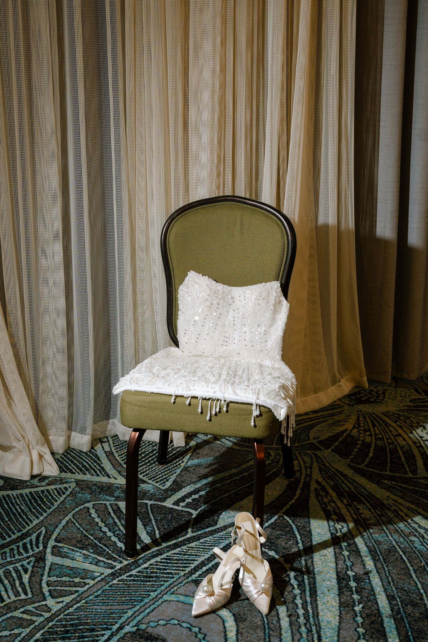 White lace wedding dress laid over an olive green chair with bridal shoes on the floor.