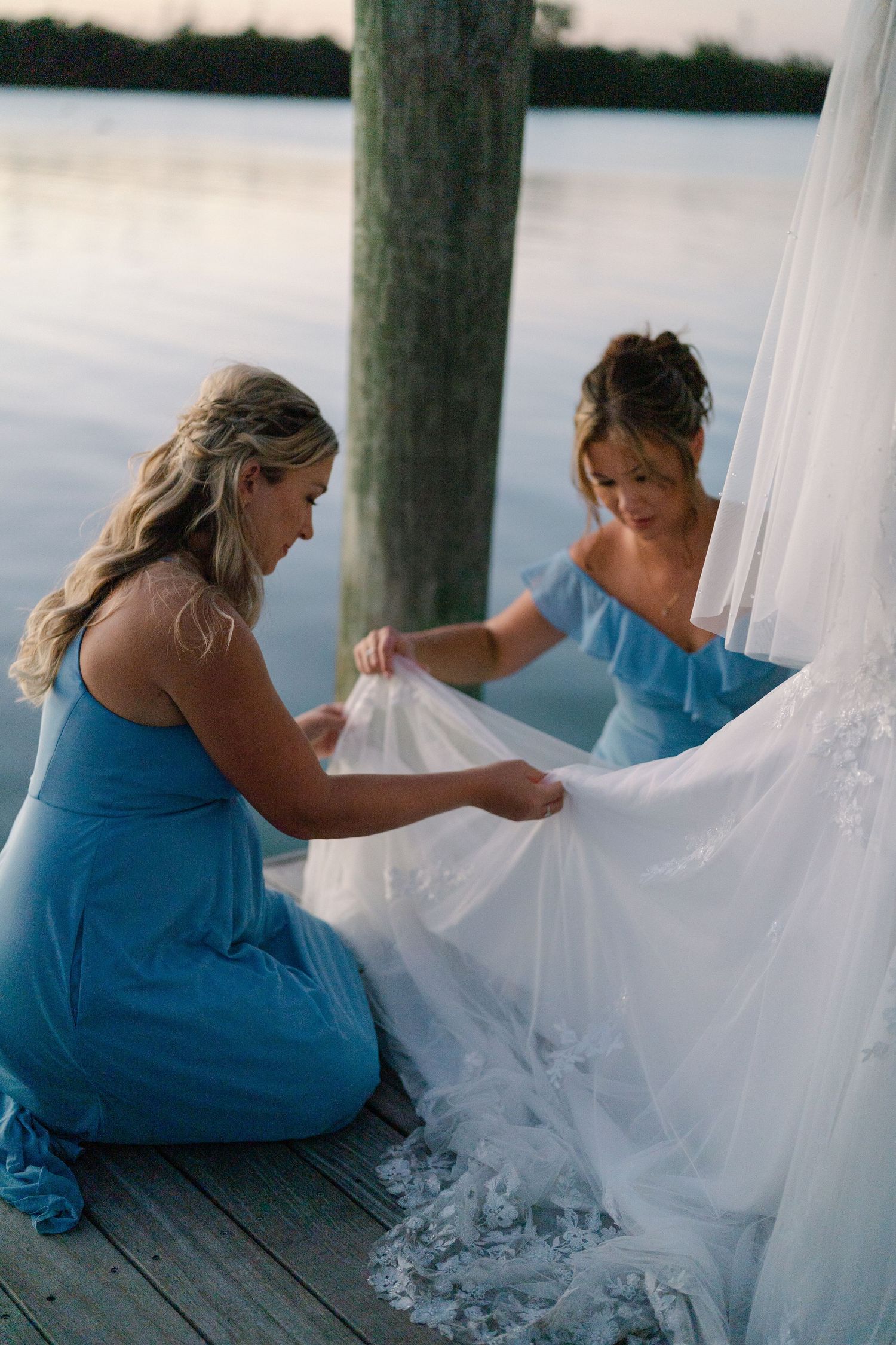 A bridesmaid in a blue dress helps arrange the train of a white wedding gown on a dock.