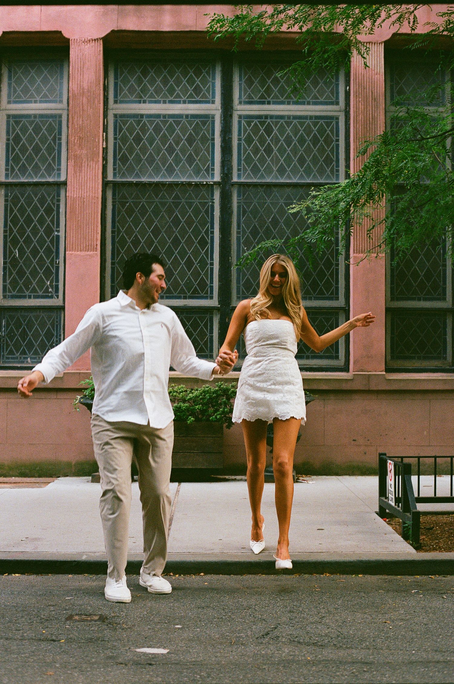 A couple in white outfits hold hands and walk joyfully on a city sidewalk beside a brick building with large windows.
