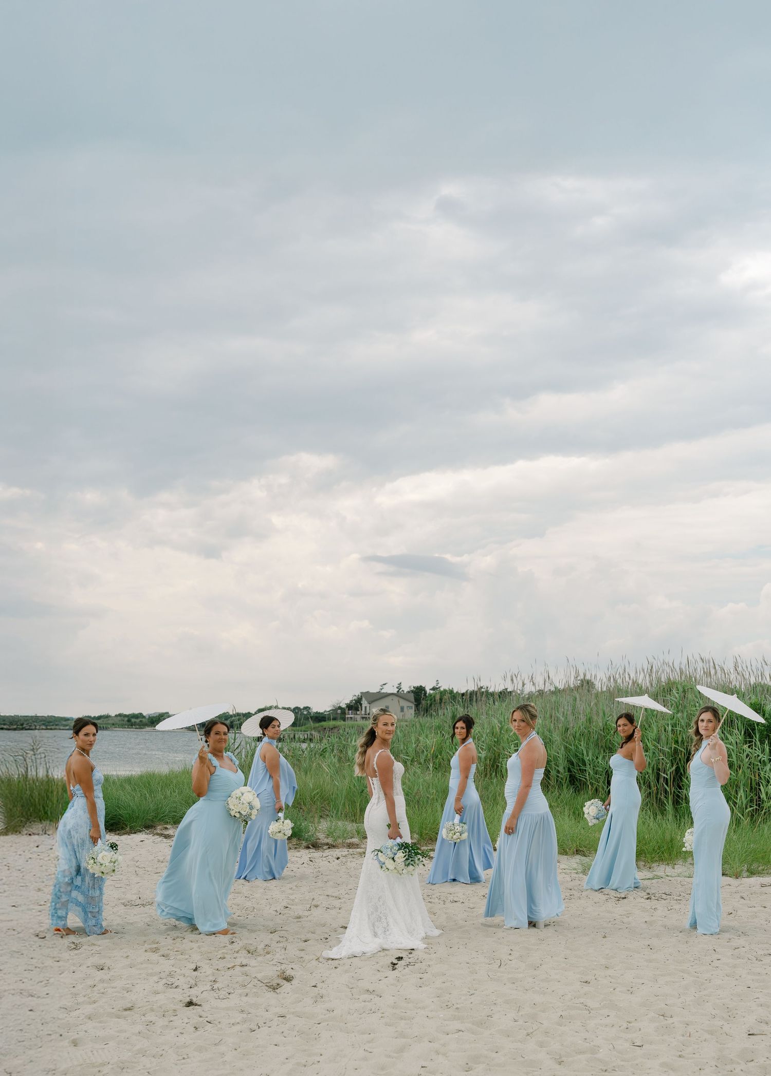 Bridesmaids in light blue gowns holding bouquets and parasols on a sandy beach with cloudy skies.