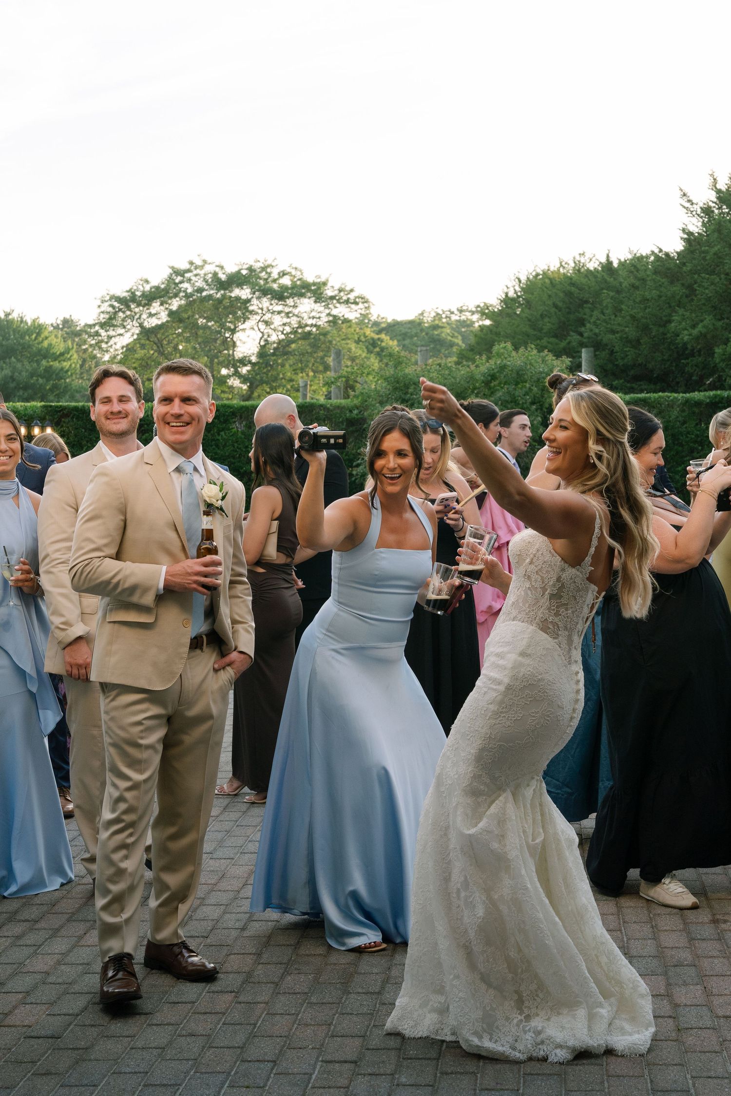 Bride in lace gown and bridesmaids in blue dresses celebrate with guests during an outdoor wedding cocktail hour.