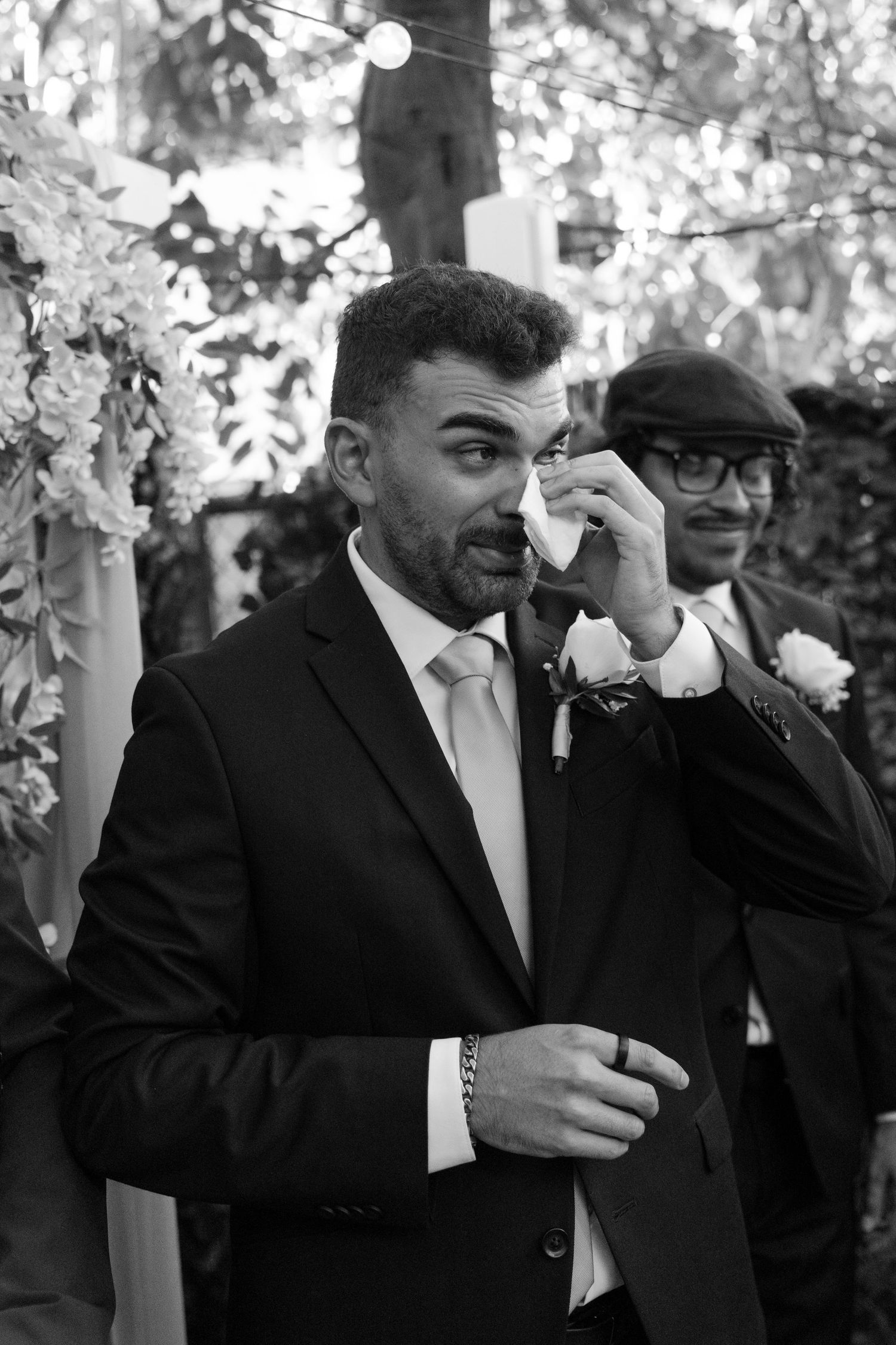 Black and white photo of emotional groom wiping tears at wedding ceremony with best man standing behind him.