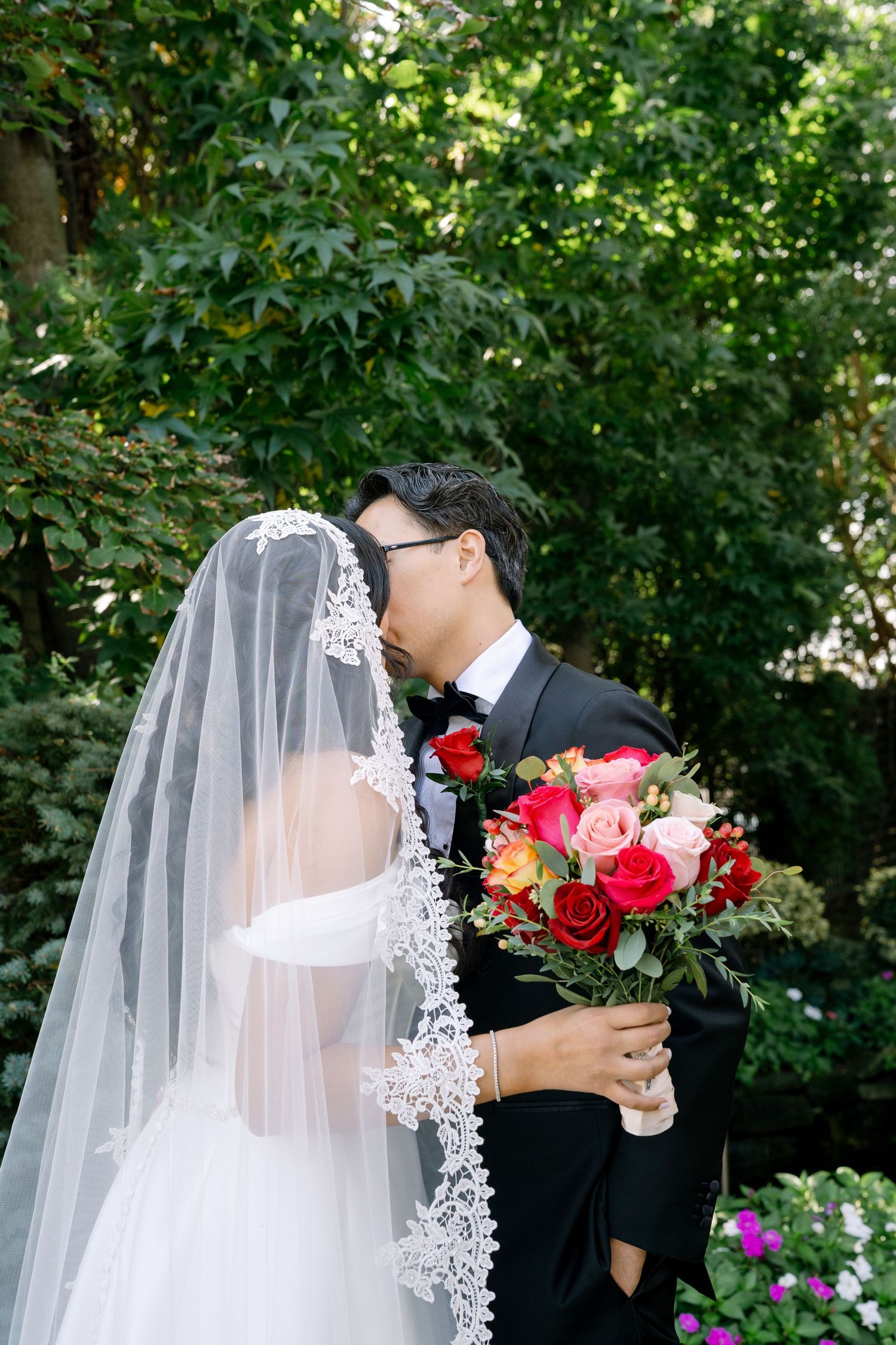 A romantic wedding moment captured in a garden setting with a flowing white veil and vibrant red bouquet.
