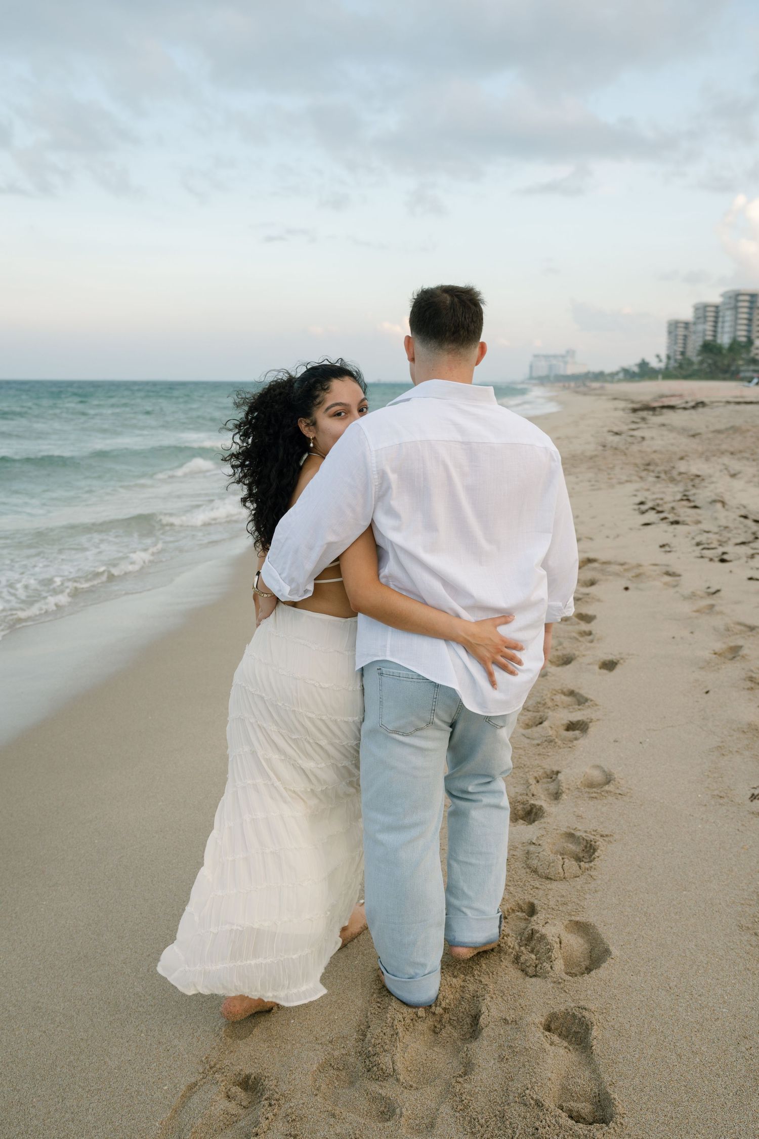 A couple in white and light blue clothing walking together along a sandy beach with ocean waves in the background.
