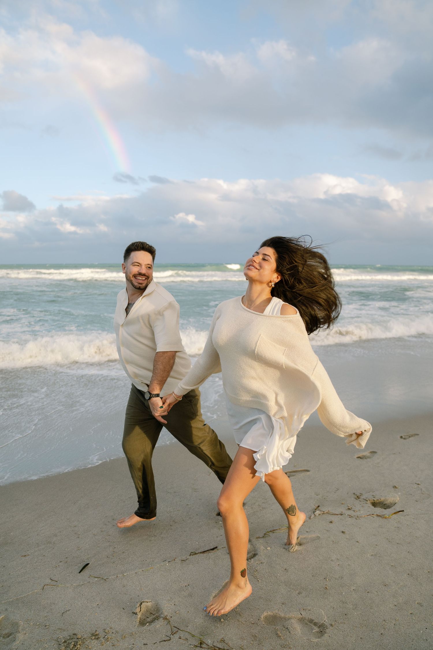 Two people dance and twirl barefoot on a beach with ocean waves and cloudy skies in the background.