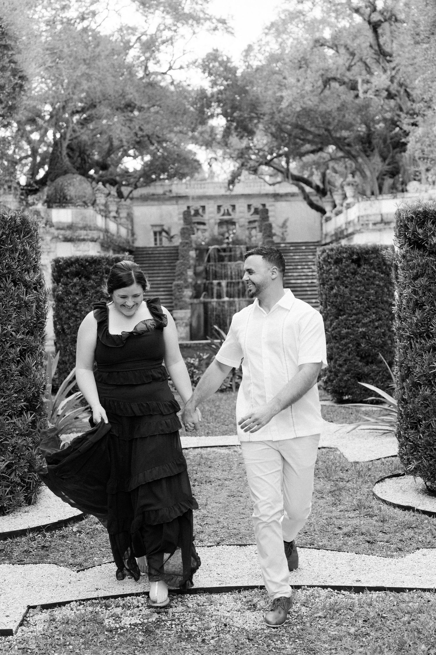 A couple walks together in a garden setting with trees and hedges, the woman wearing a black dress and man in white.