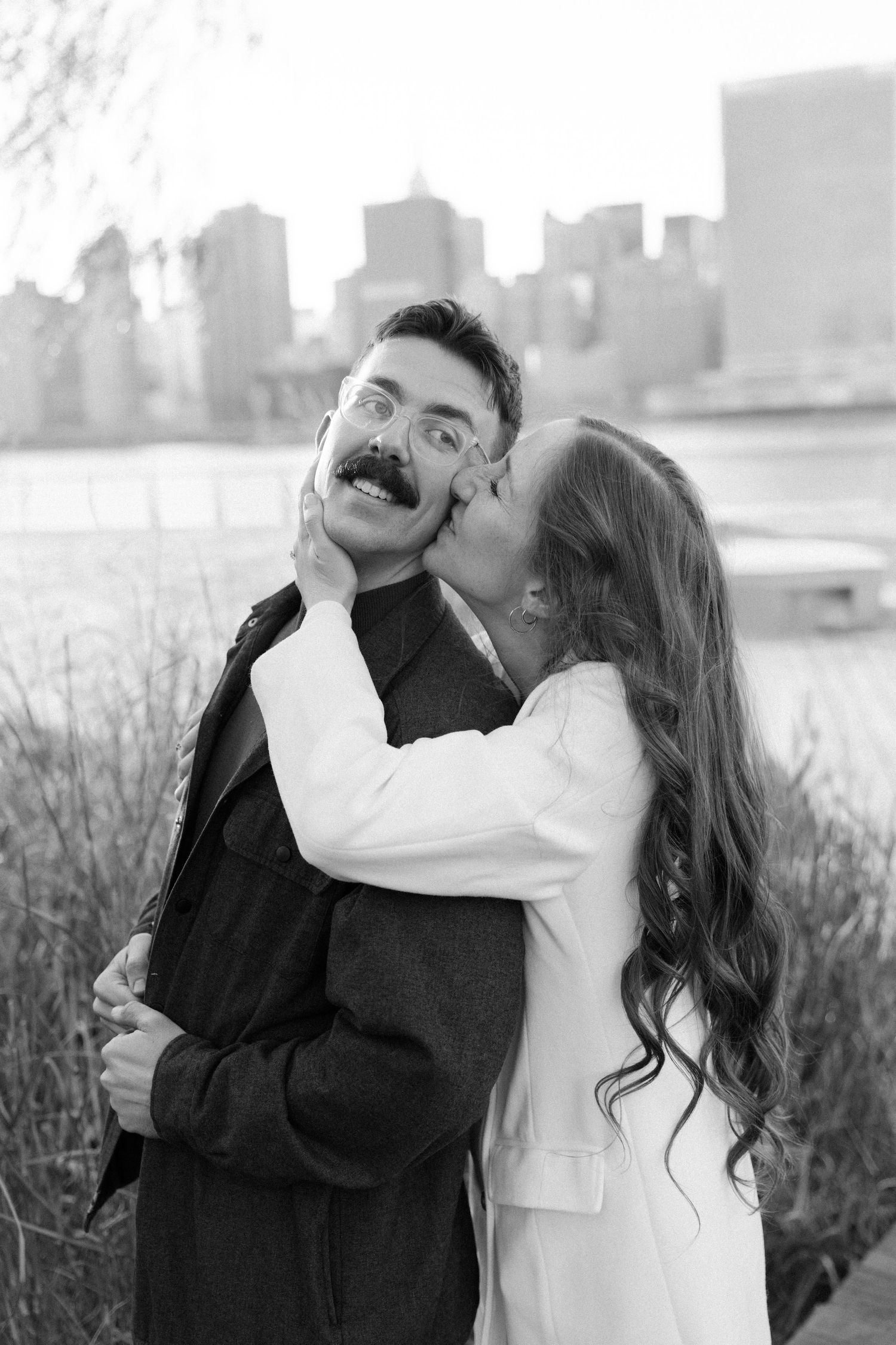 A romantic couple embraces in multiple candid black and white shots with an urban waterfront backdrop.