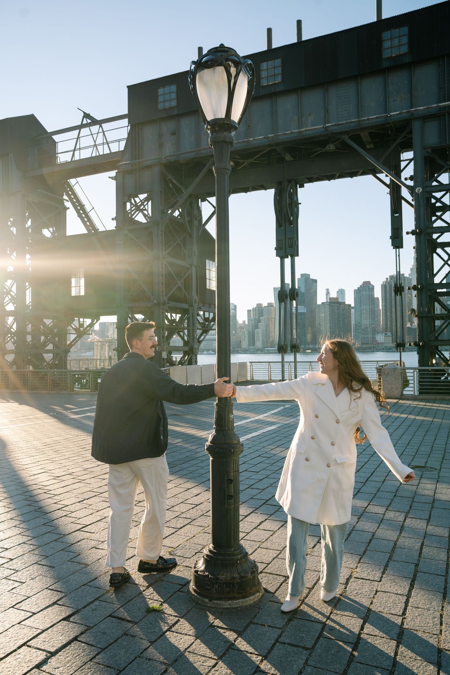 Two people dance in silhouette on a pier with sunlight streaming through industrial structures.