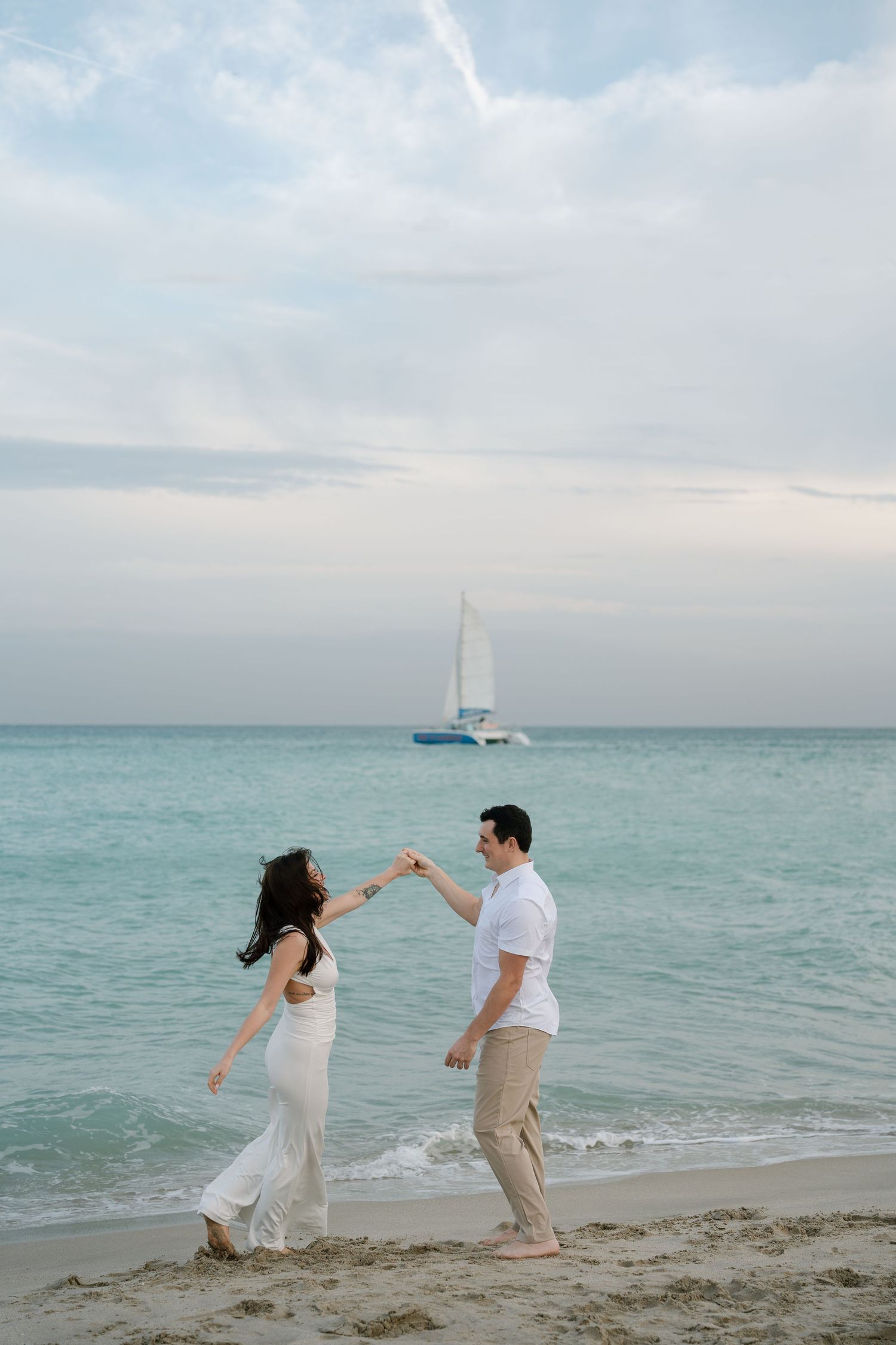 A couple dances playfully together on a beach with turquoise waters and a sailboat in the background.