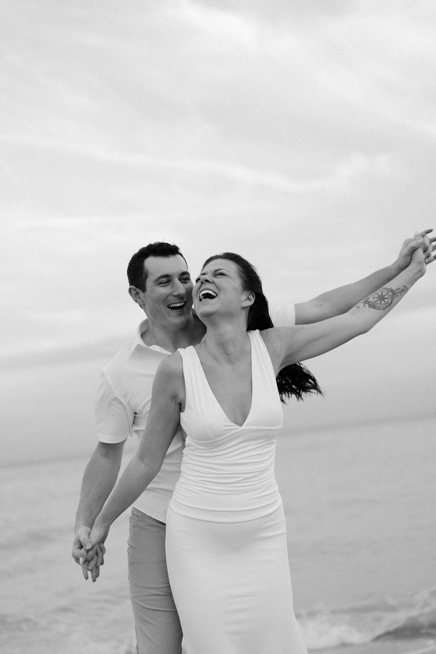 A joyful couple in white outfits spreads their arms while laughing together on a cloudy beach.