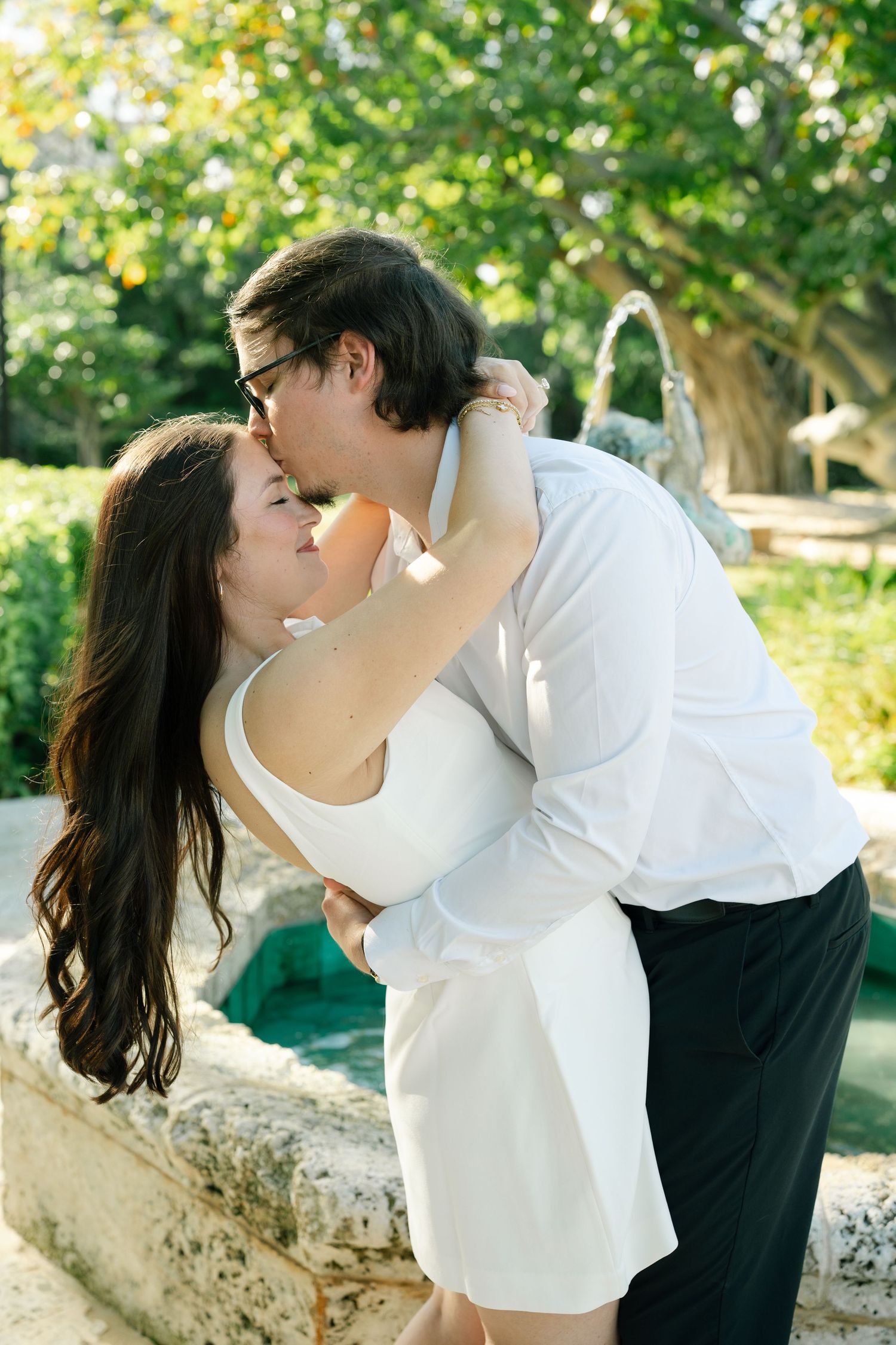 A romantic couple shares tender kisses in a sunlit outdoor garden setting with lush greenery in the background.