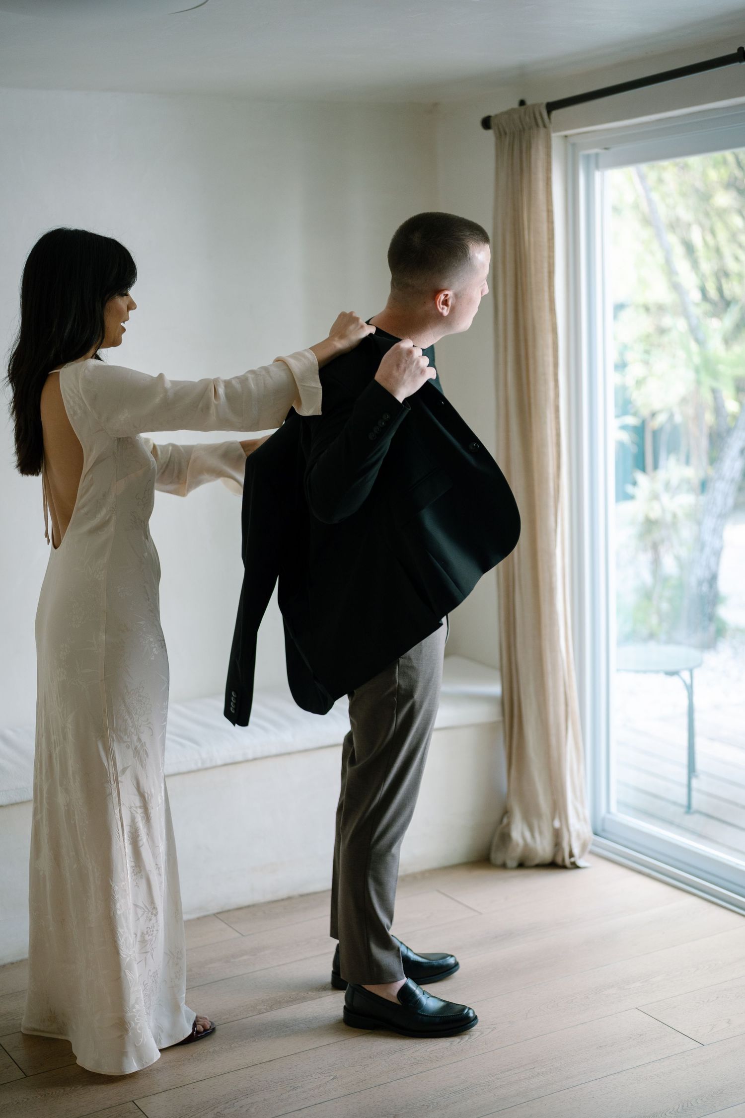 A tender moment of wedding preparation as a dress is adjusted in a sunlit room.