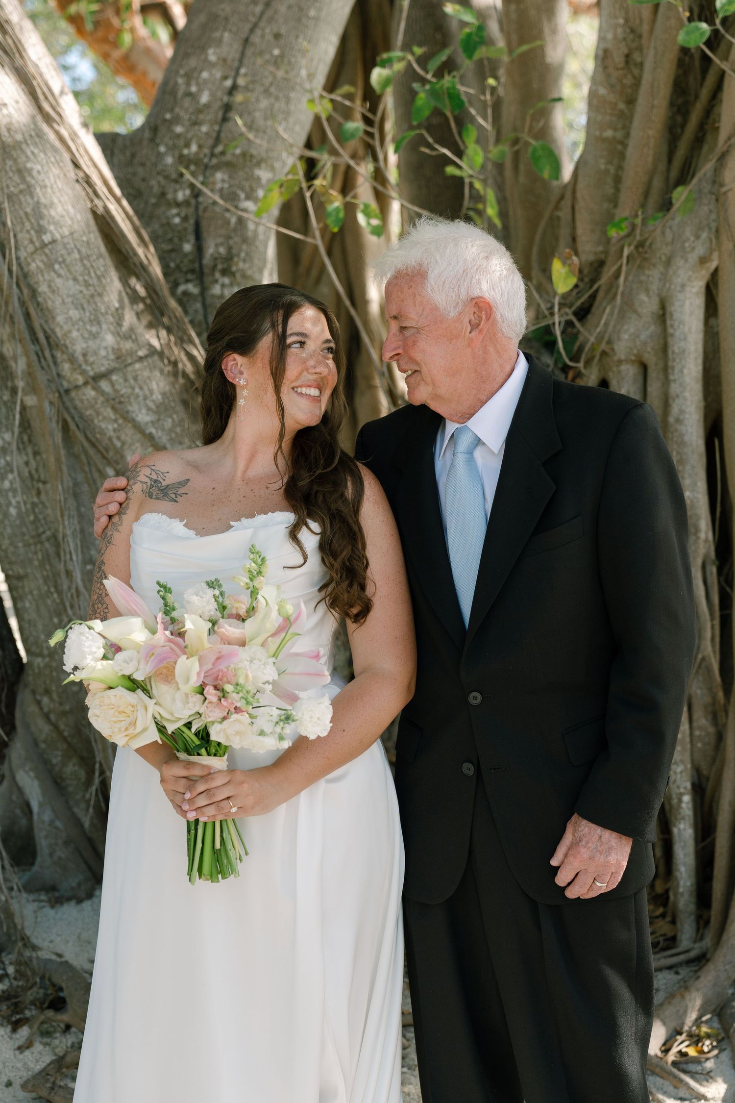 A bride and her elderly father walk together under the shade of trees.