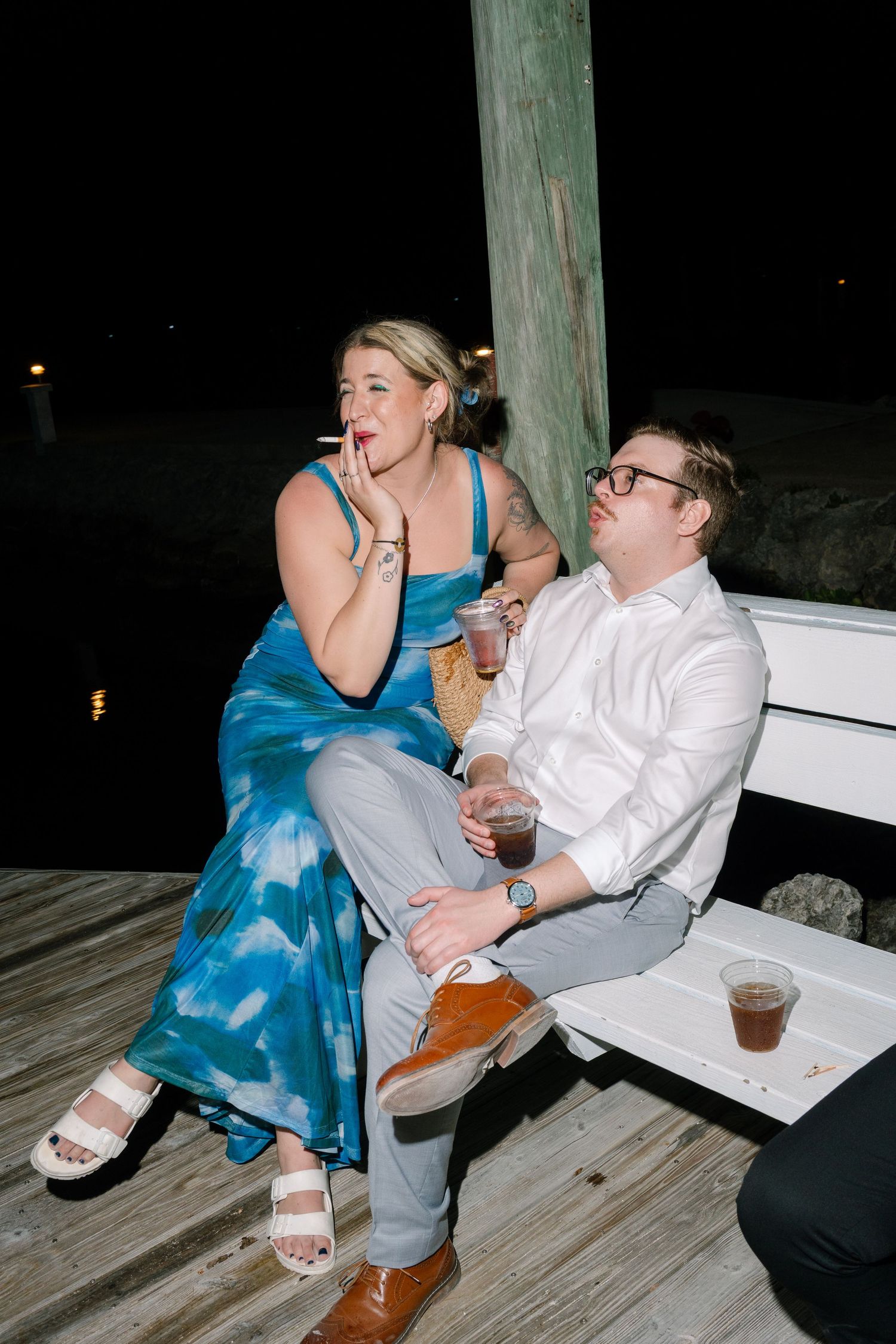 Two people, a woman in a blue dress and a man in a white shirt, sitting together and laughing on an outdoor deck at night.
