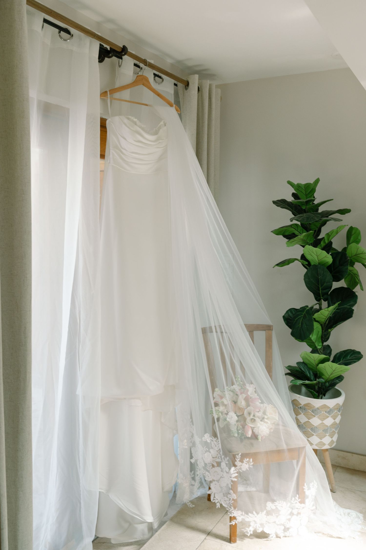 A white wedding dress hanging on a clothing rack in a room with a potted plant.