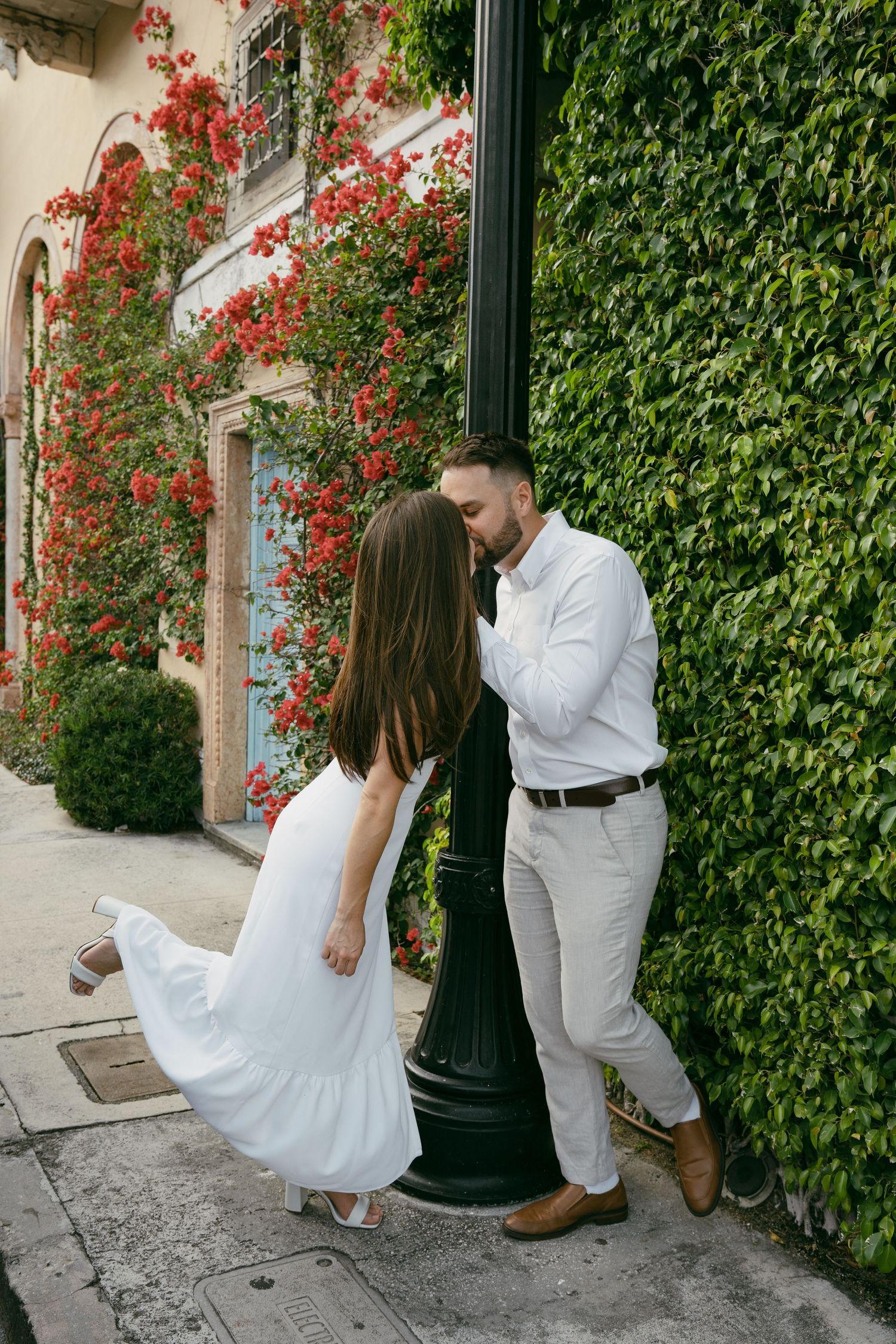 A couple in white outfits share a romantic moment against an ivy wall with blooming red flowers.