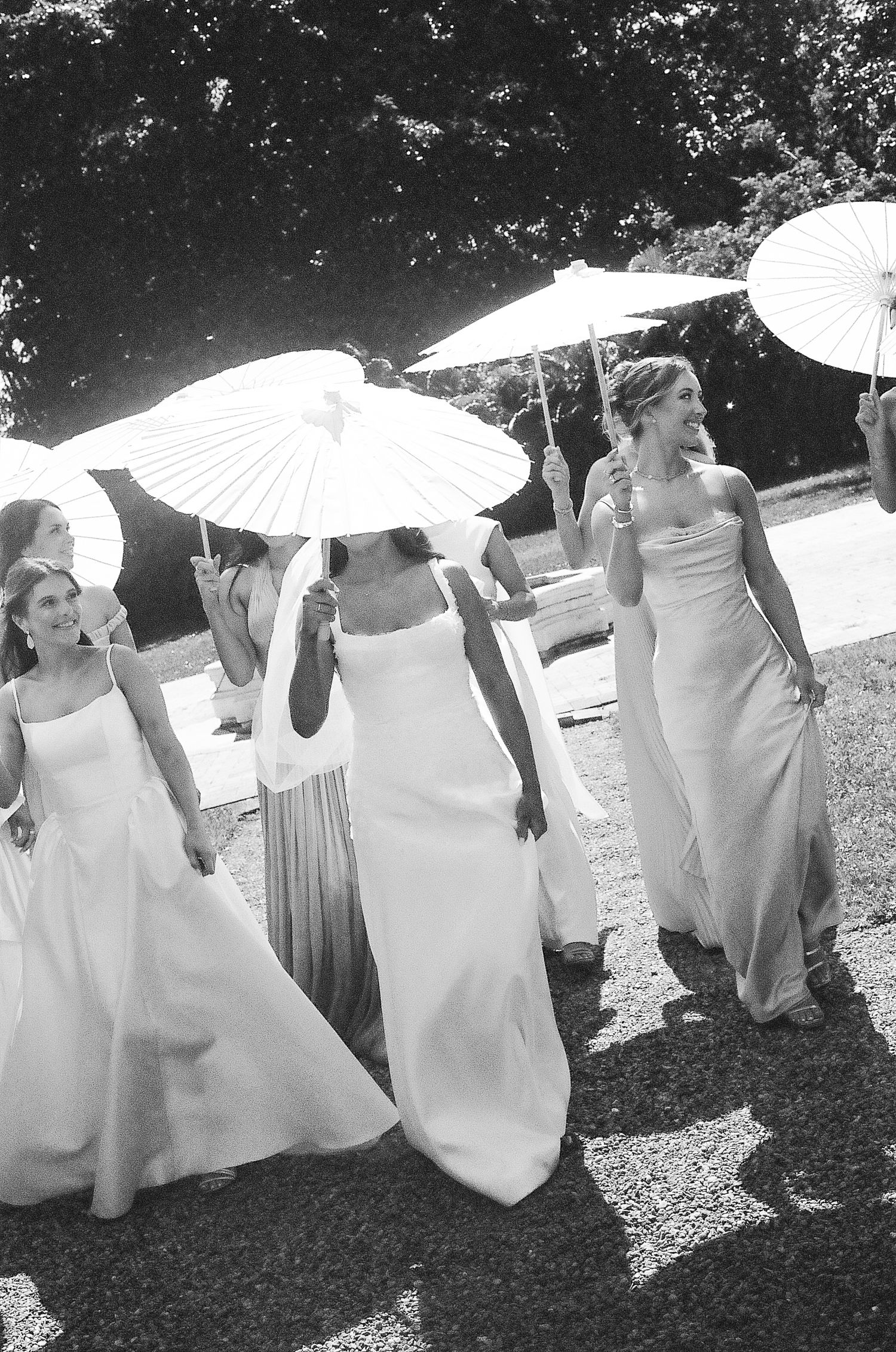 Bridesmaids in white gowns walk outdoors holding white parasol umbrellas at a wedding celebration.