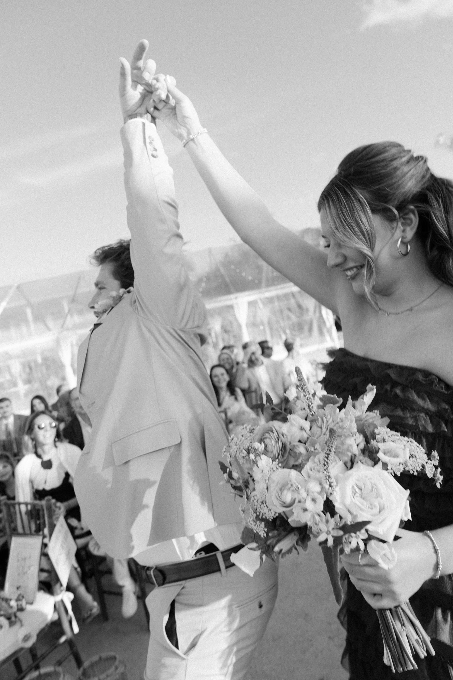 Black and white photo of joyful wedding celebration with guests raising hands and holding bouquets.