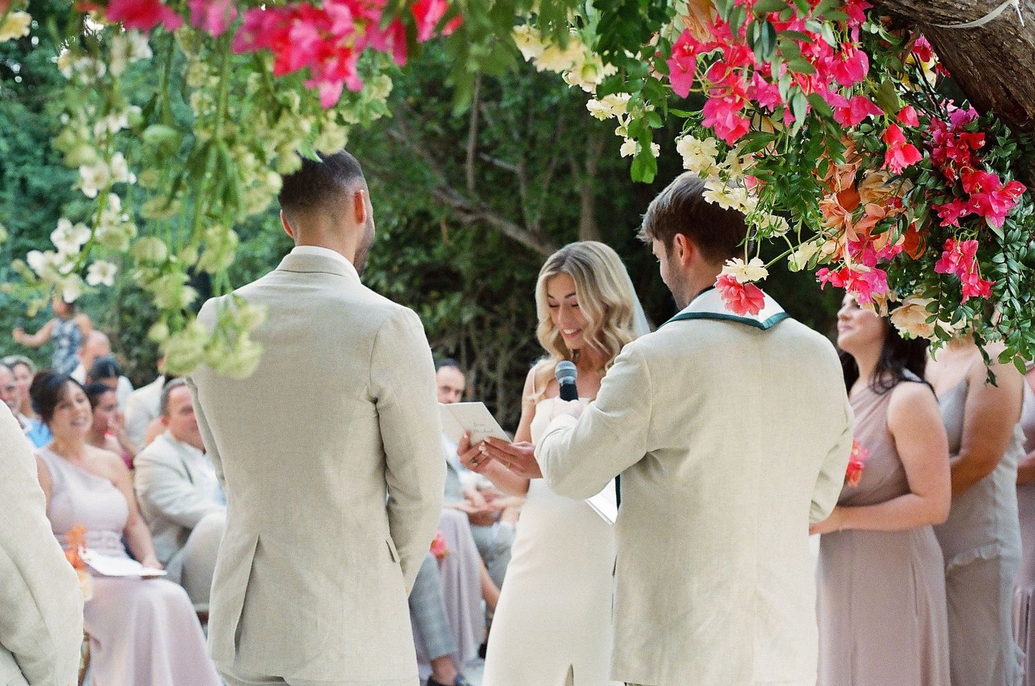 Wedding party in cream and white attire standing under vibrant pink floral arrangements.