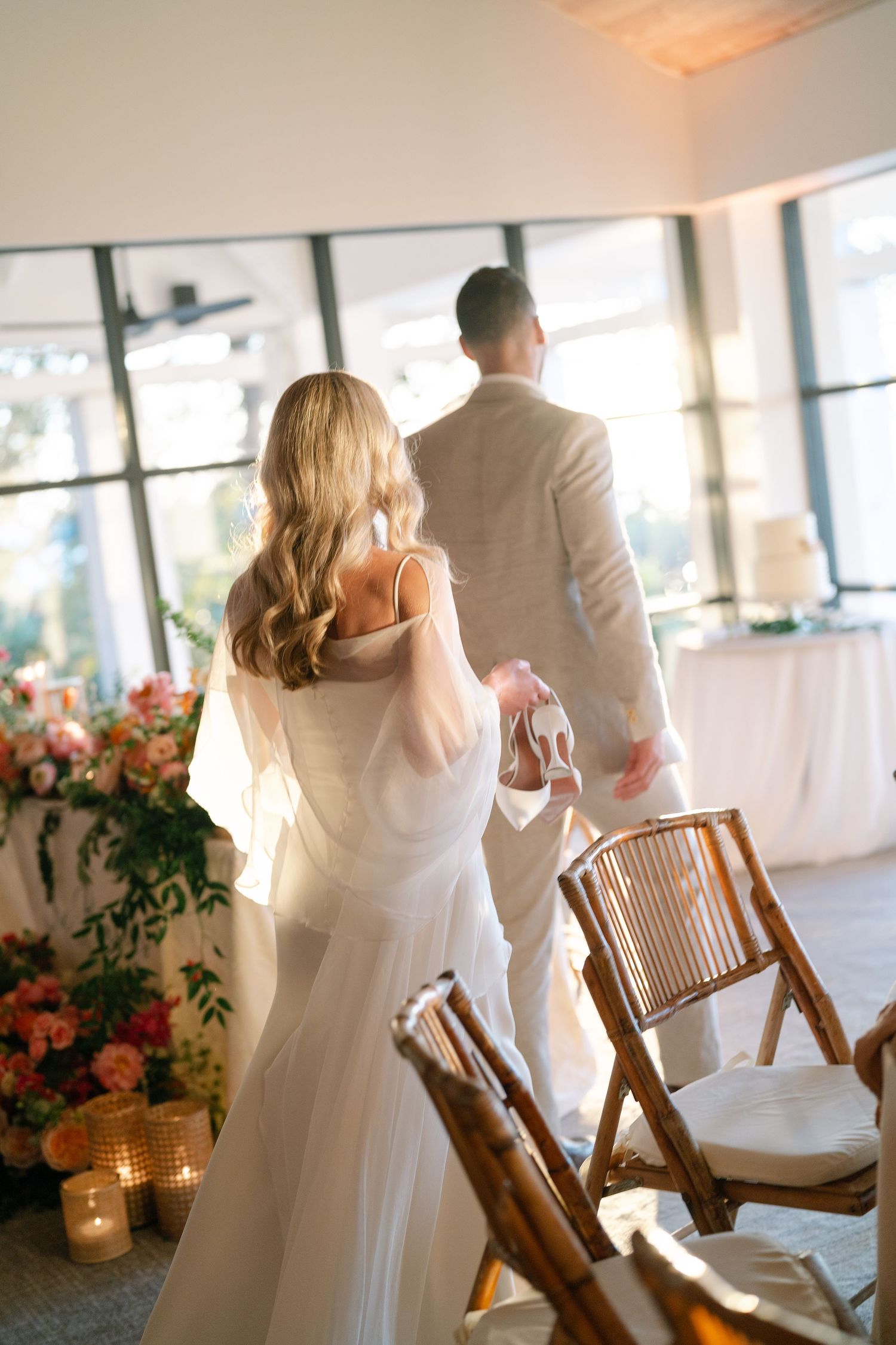 A couple in white attire dance together in a bright indoor venue decorated with coral floral arrangements.