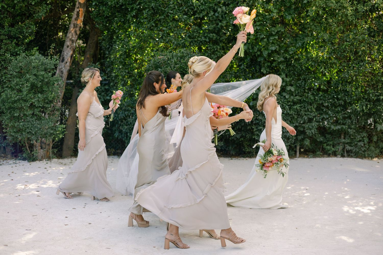 Wedding party members in light colored dresses dancing joyfully on a white floor outdoors.