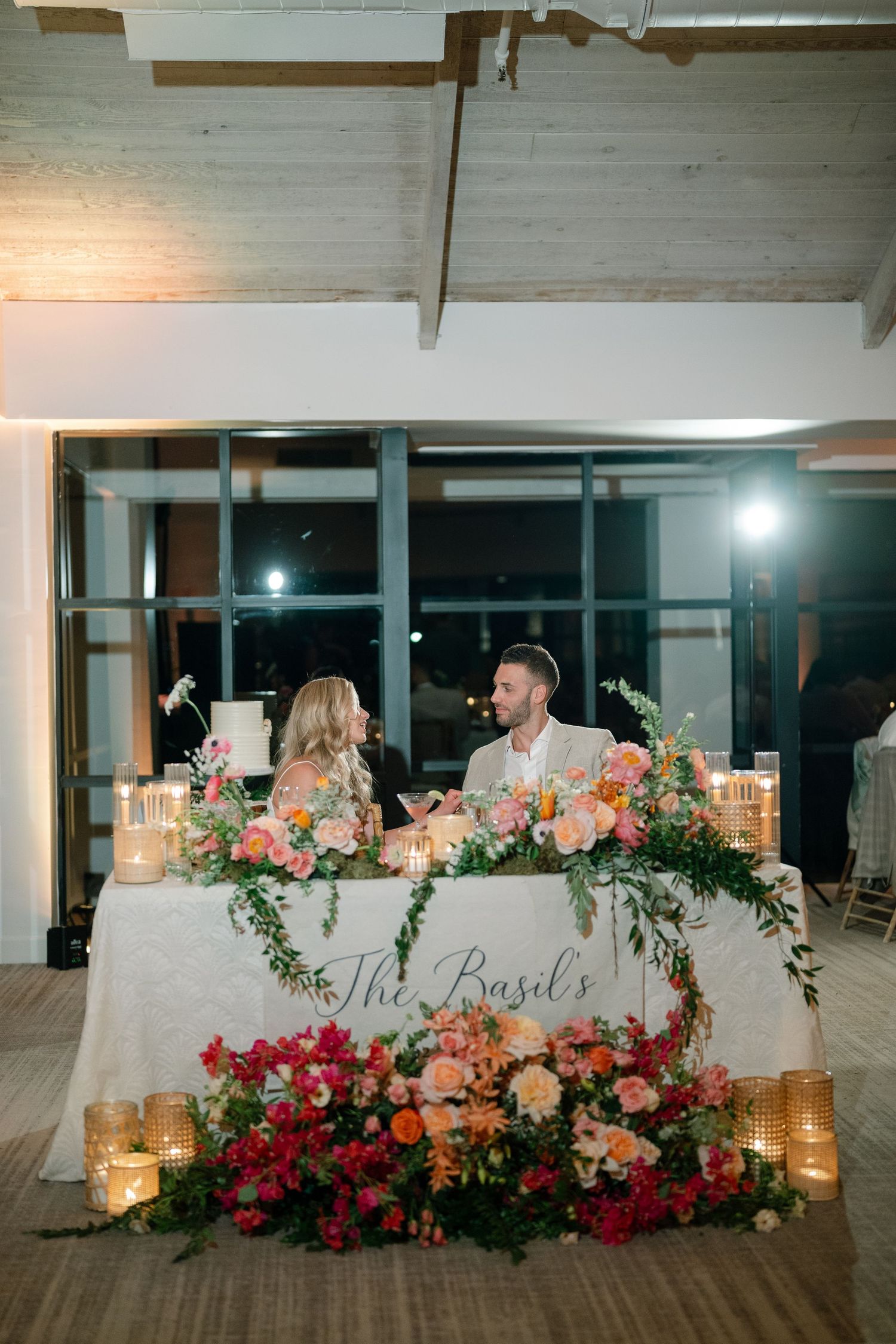 Wedding reception head table decorated with pink and peach floral arrangements and glowing candles.