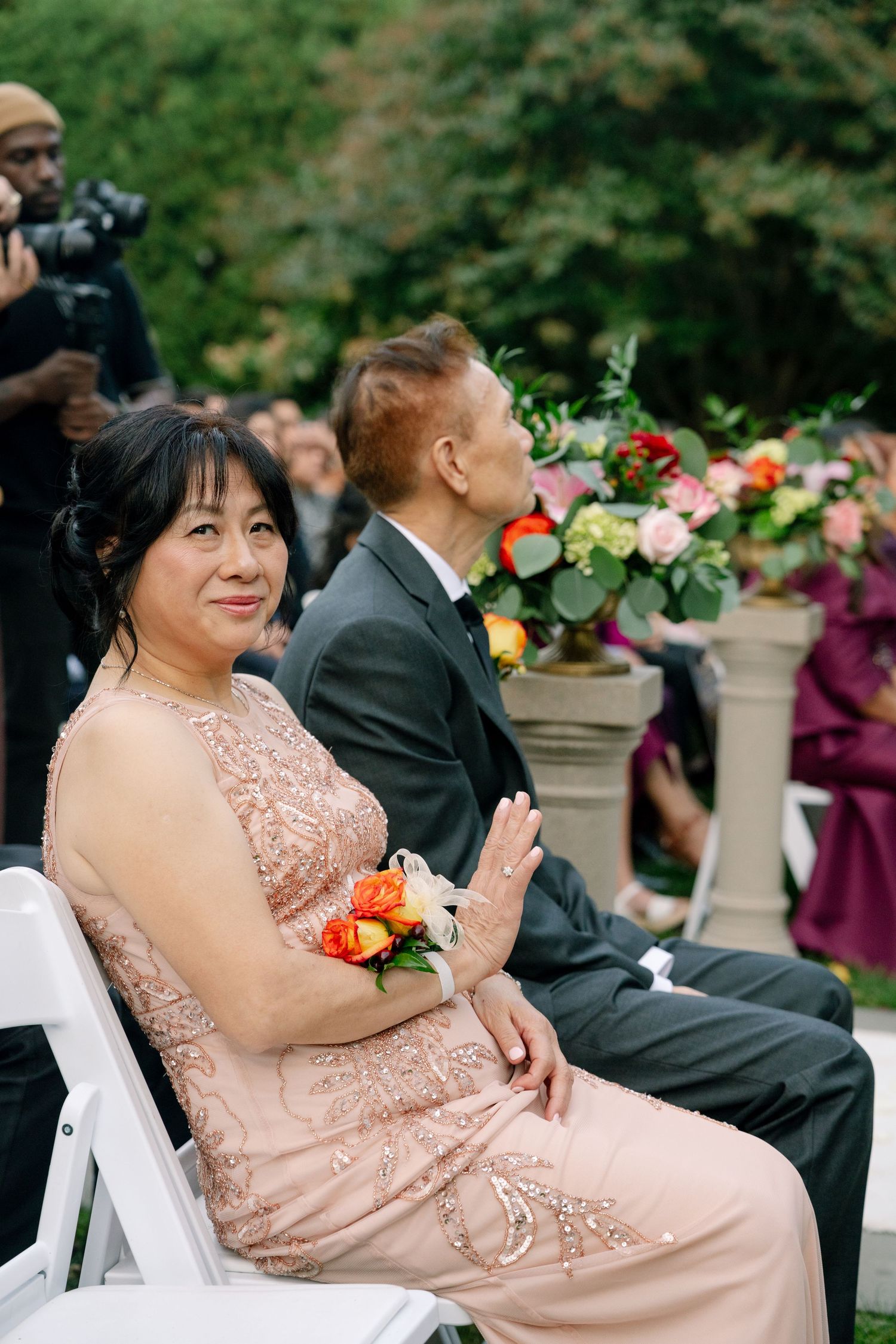 Wedding guests in formal attire seated at outdoor ceremony with floral arrangements.