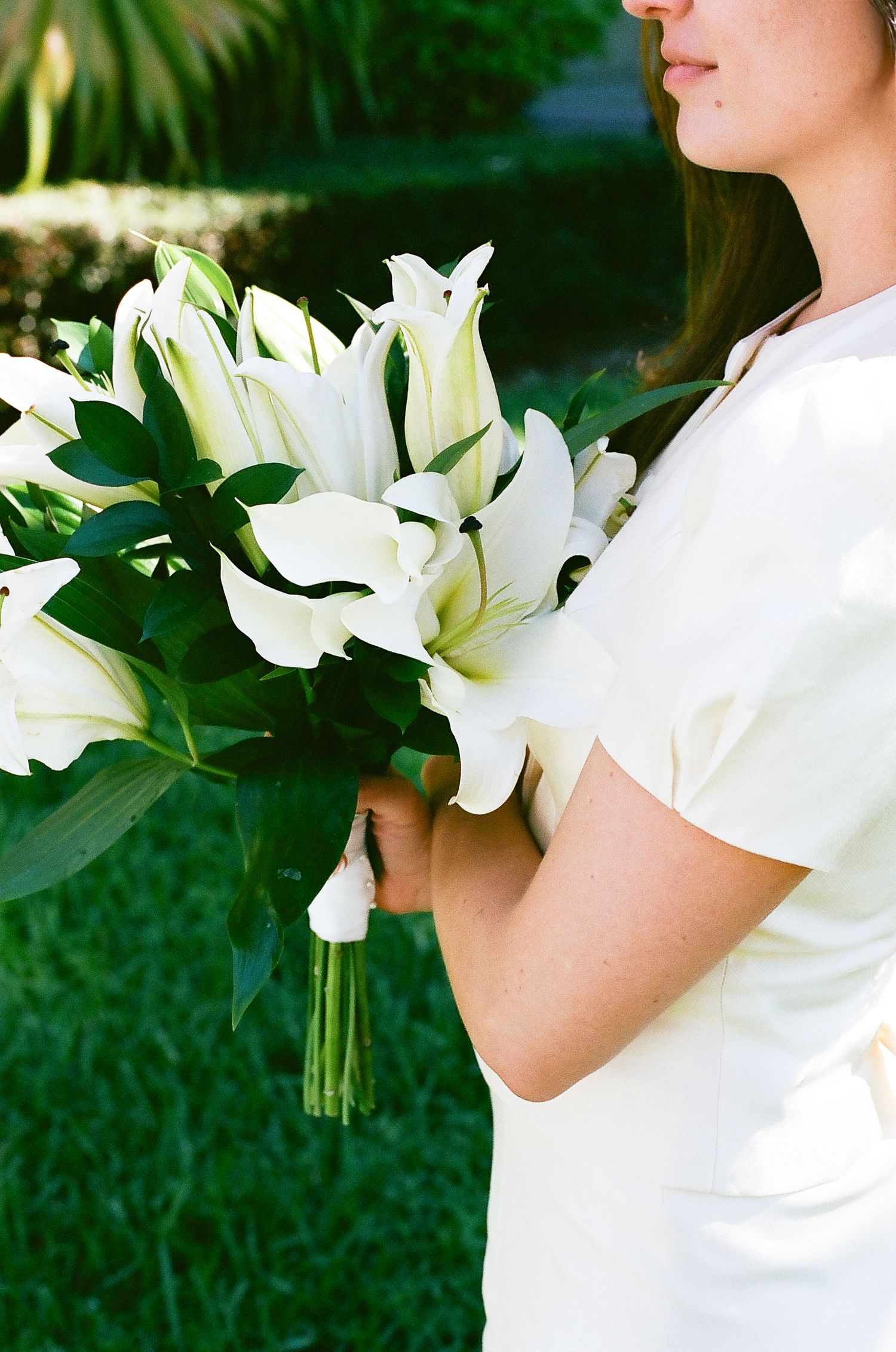 White lily bouquet held against white dress in outdoor garden setting.