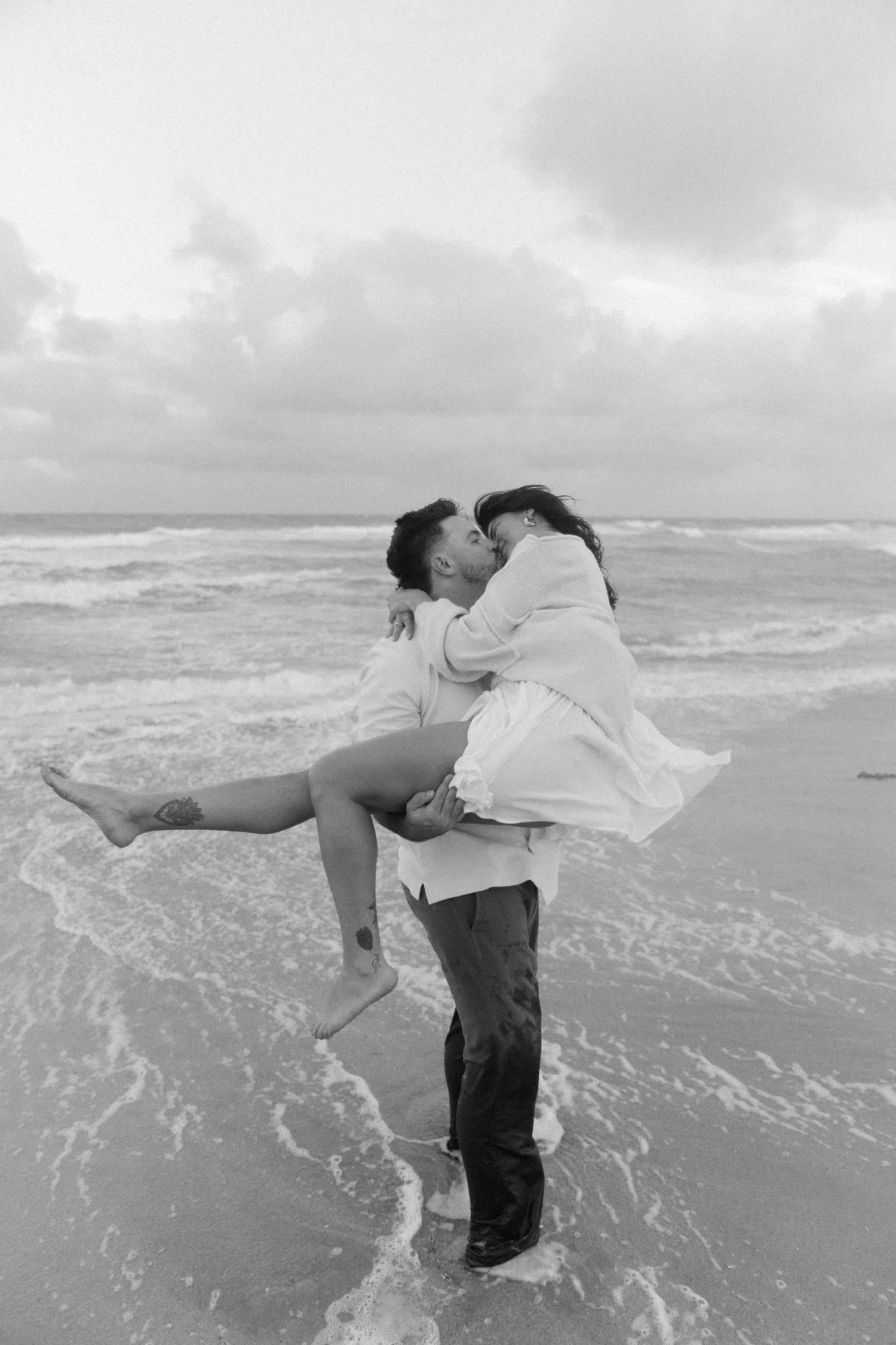 A playful sequence showing a couple dancing and spinning together at the ocean's edge in black and white.