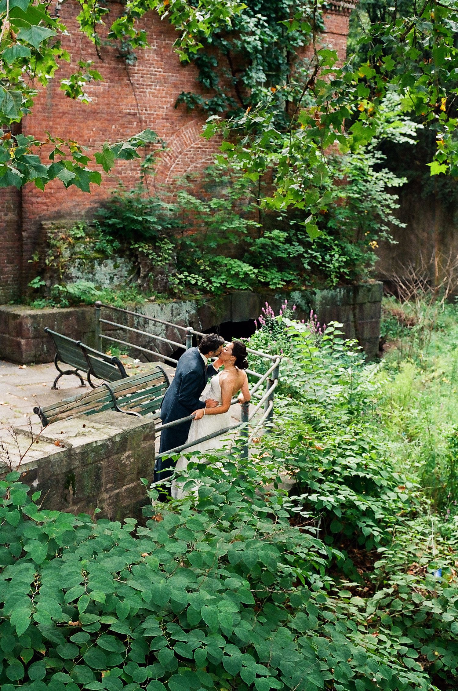 A couple shares an intimate moment on a park bench surrounded by lush greenery and a brick wall backdrop.