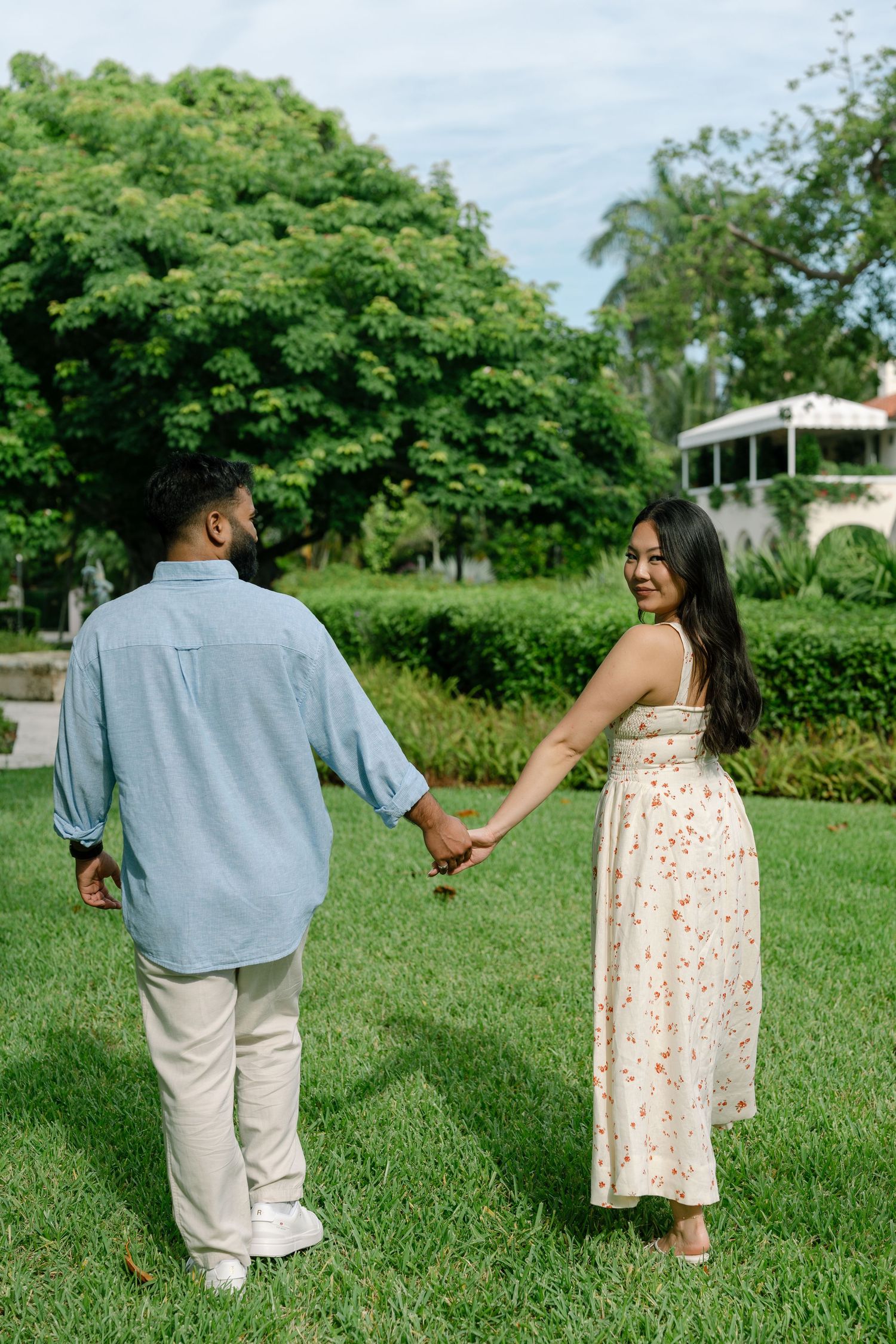 Couple holding hands in a lush green park, woman in floral dress smiling back at camera.