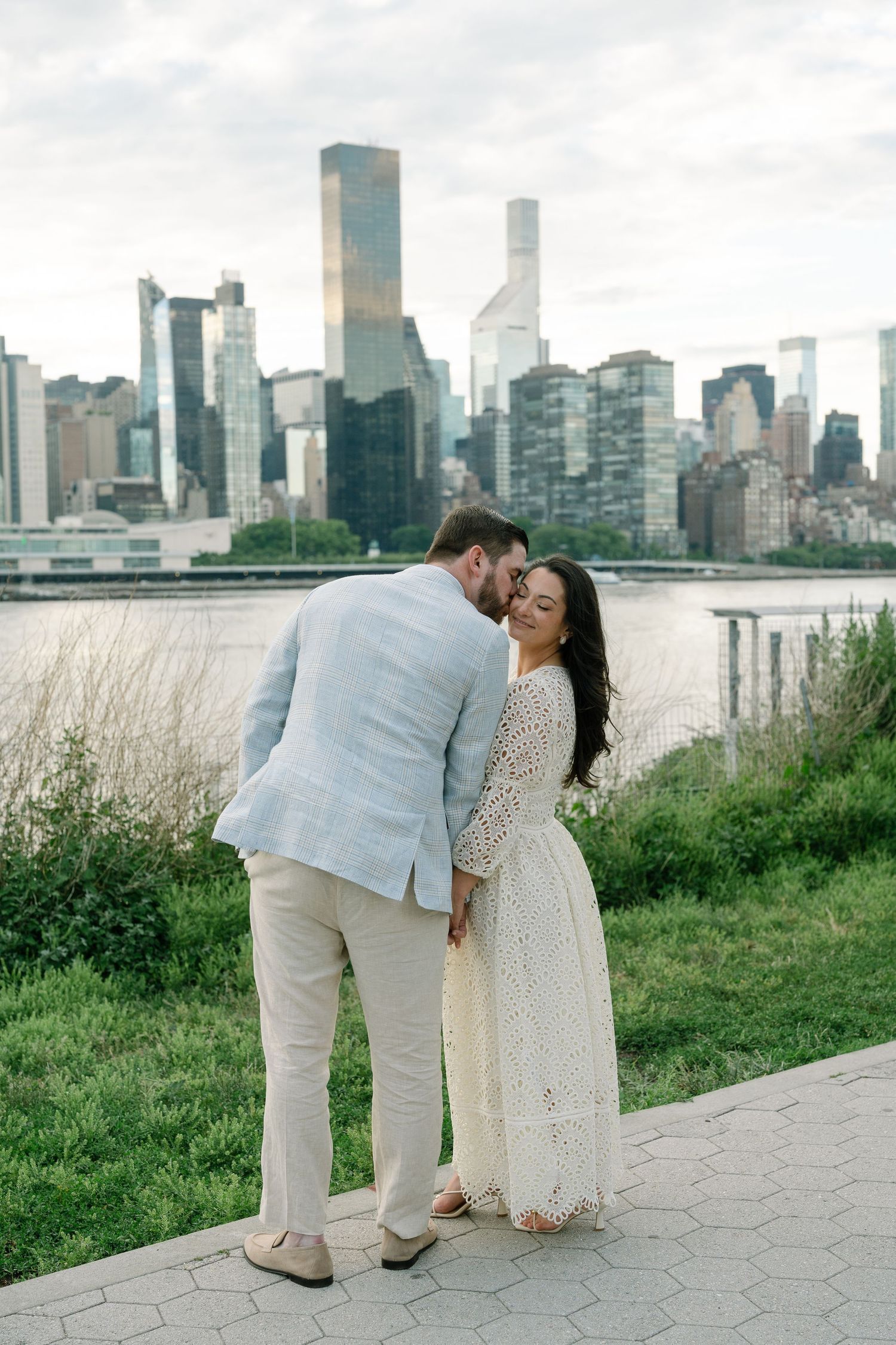 Couple sharing a romantic moment with NYC skyline and river backdrop, man in blue blazer kissing woman in white lace dress.